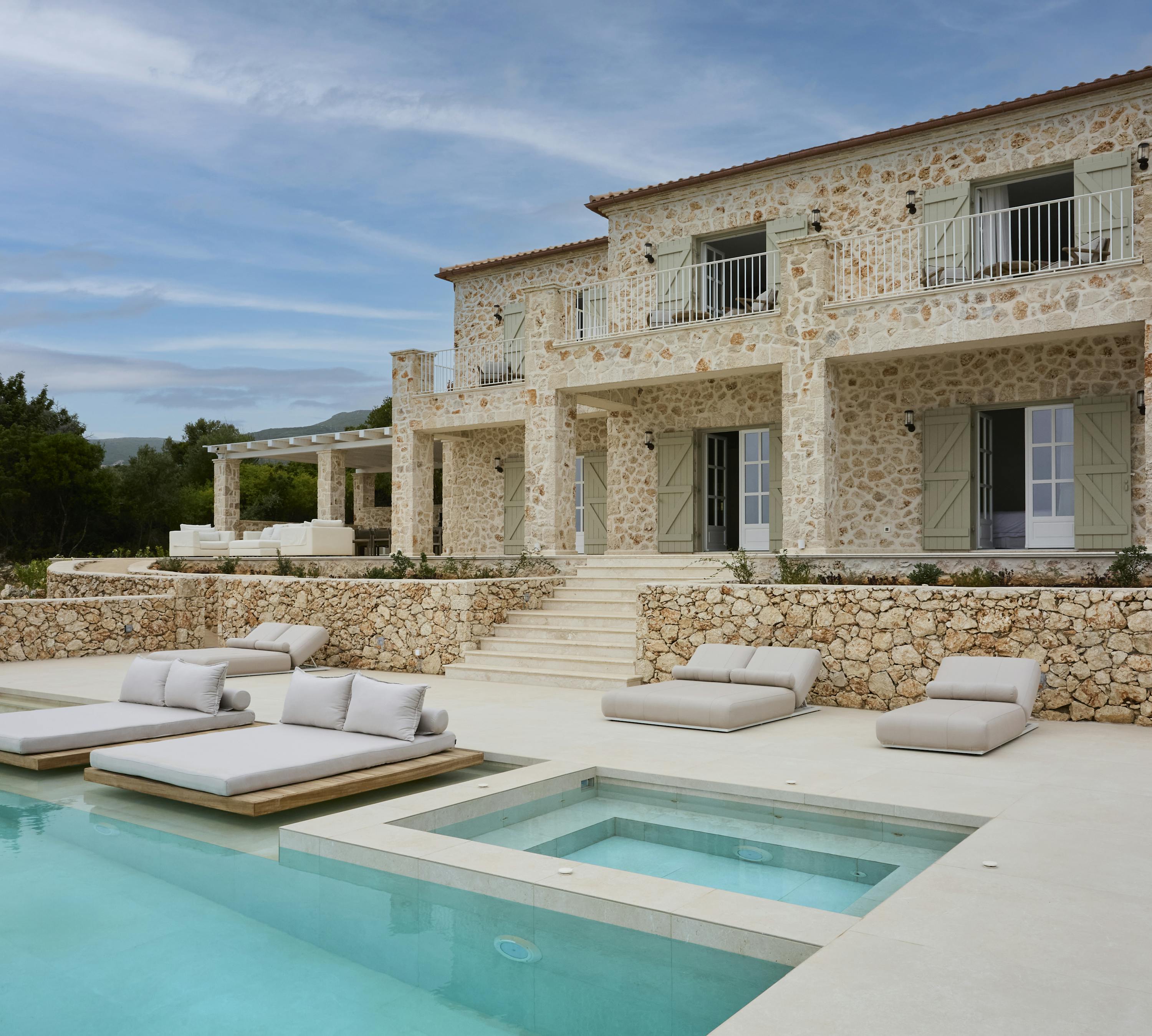 The villa's impressive stone facade showcases traditional Mediterranean architecture with sage green shutters and terracotta roof tiles. Multiple levels of terracing lead down to the infinity pool area with its luxury outdoor furniture.
