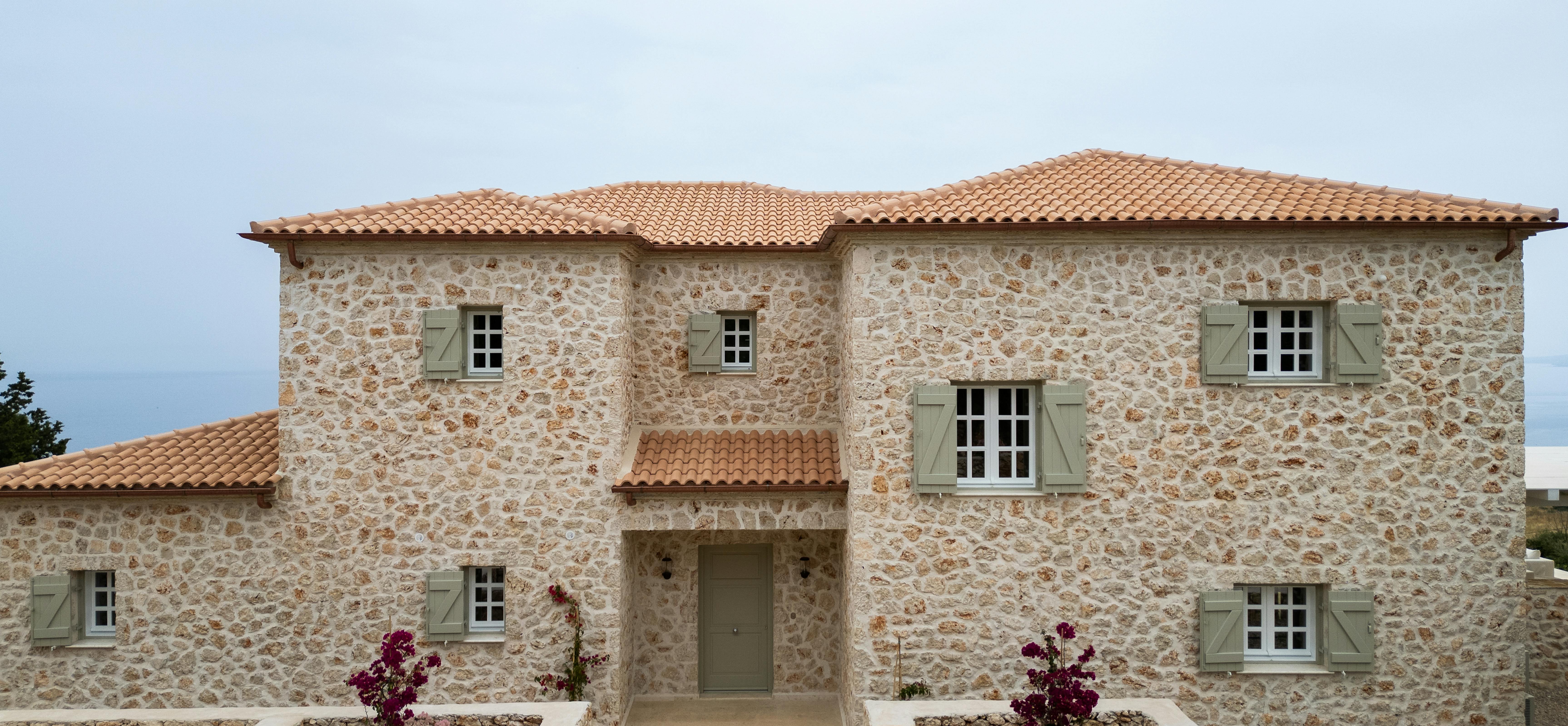 The villa's front facade showcases traditional stone construction with green wooden shutters and terracotta roof tiles. Landscaped entrance gardens with purple flowers frame the symmetrical two-storey structure.