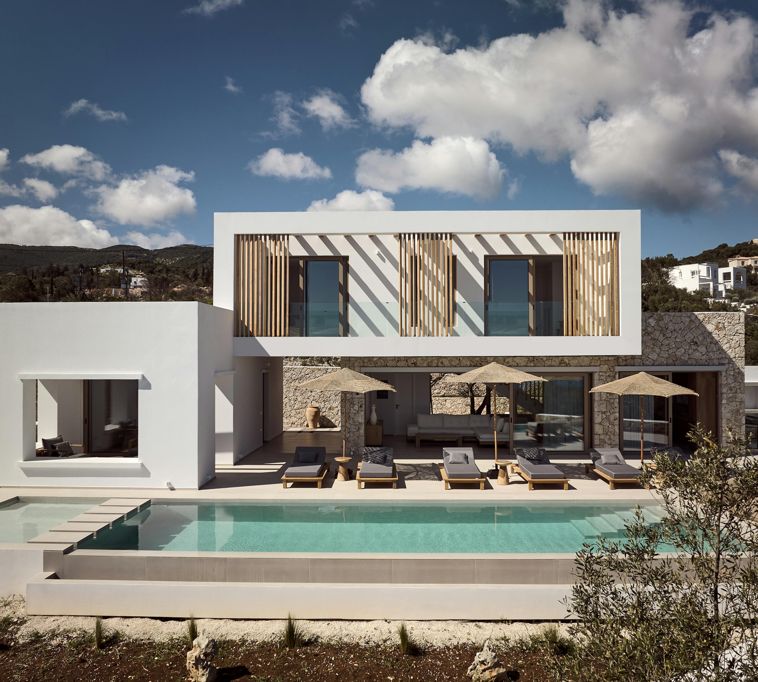 Modern two-story villa with wooden shutters, infinity pool, and dramatic hillside Mediterranean setting.