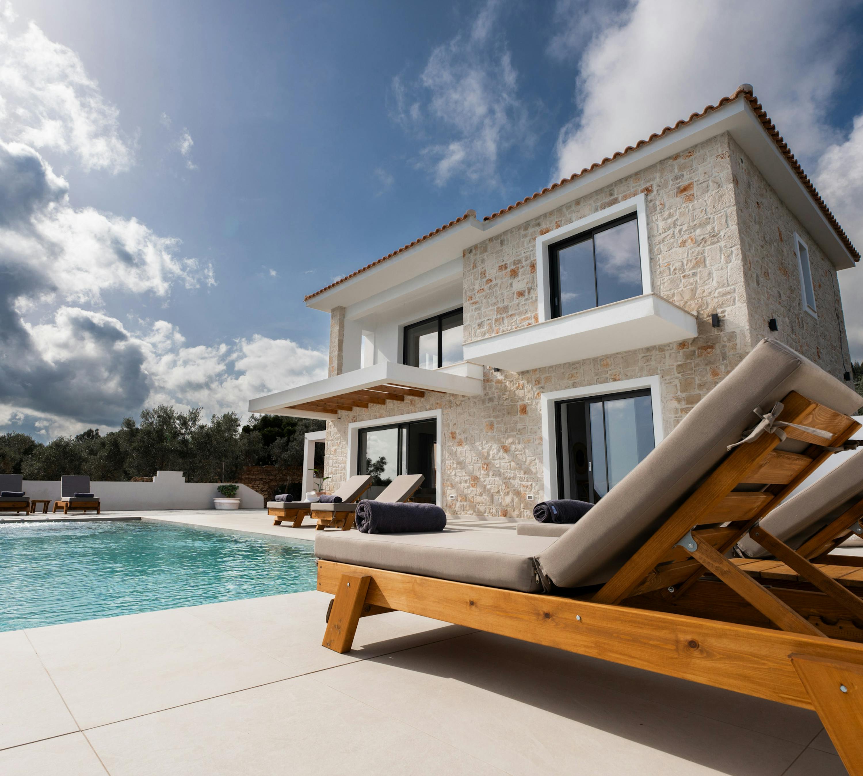 A modern Mediterranean villa with natural stone walls overlooks a swimming pool with timber sun loungers arranged on the deck. The two-storey home features contemporary design elements against a dramatic cloudy sky.