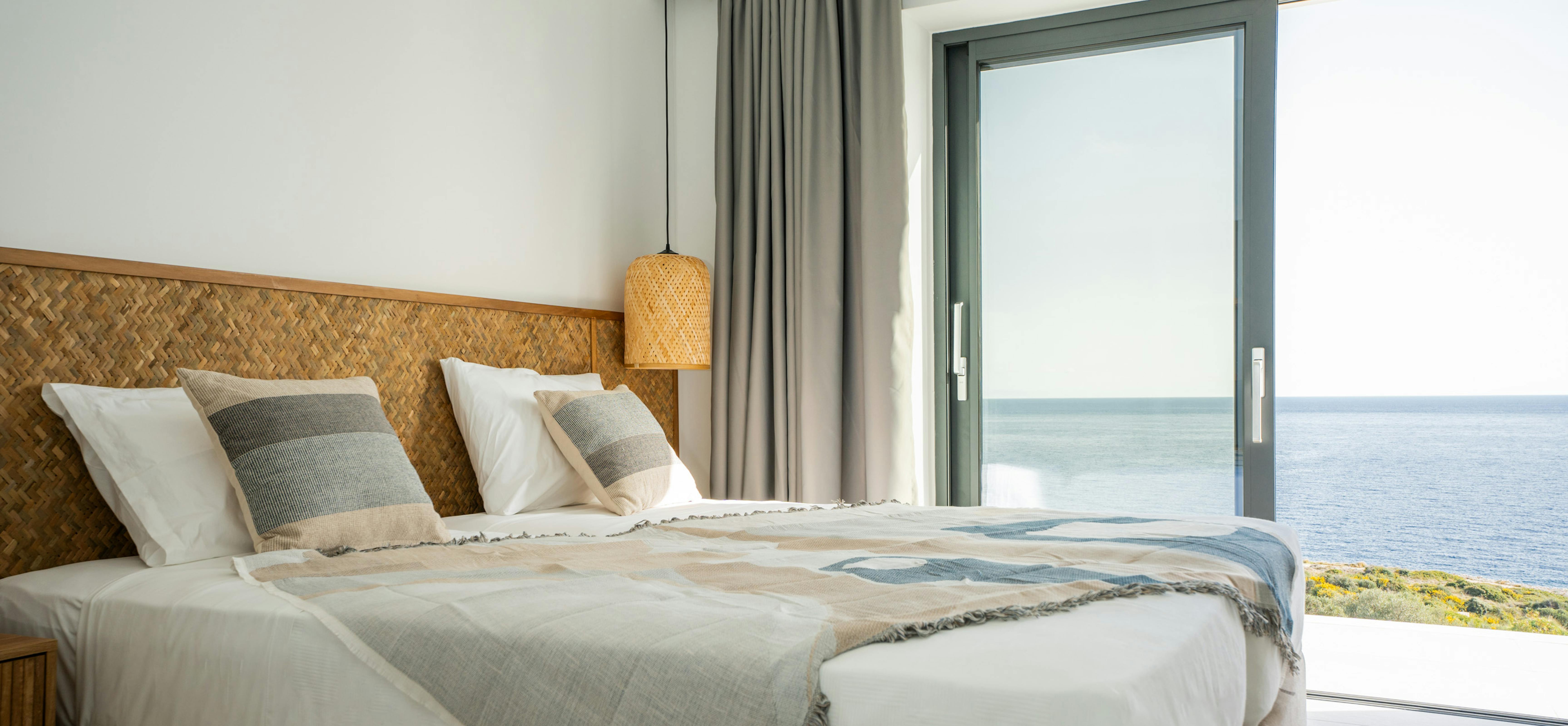 A bedroom showcases a woven rattan headboard with white bedding and striped accent cushions. Floor-to-ceiling windows provide natural light whilst a wicker pendant light adds textural interest to the space.