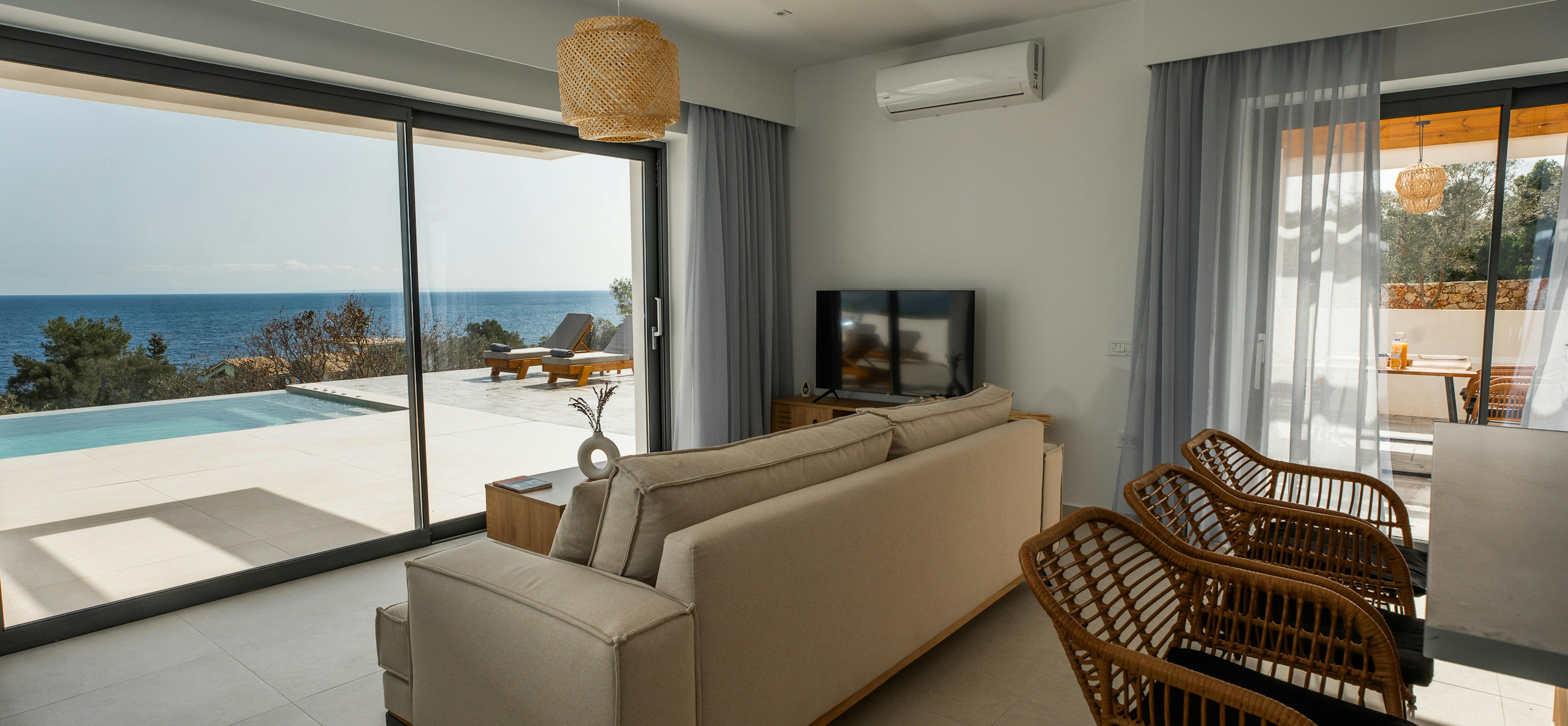 A contemporary living area features neutral seating and rattan accent chairs overlooking the pool and Mediterranean beyond. A woven pendant light and natural textures create warmth in the minimalist space.