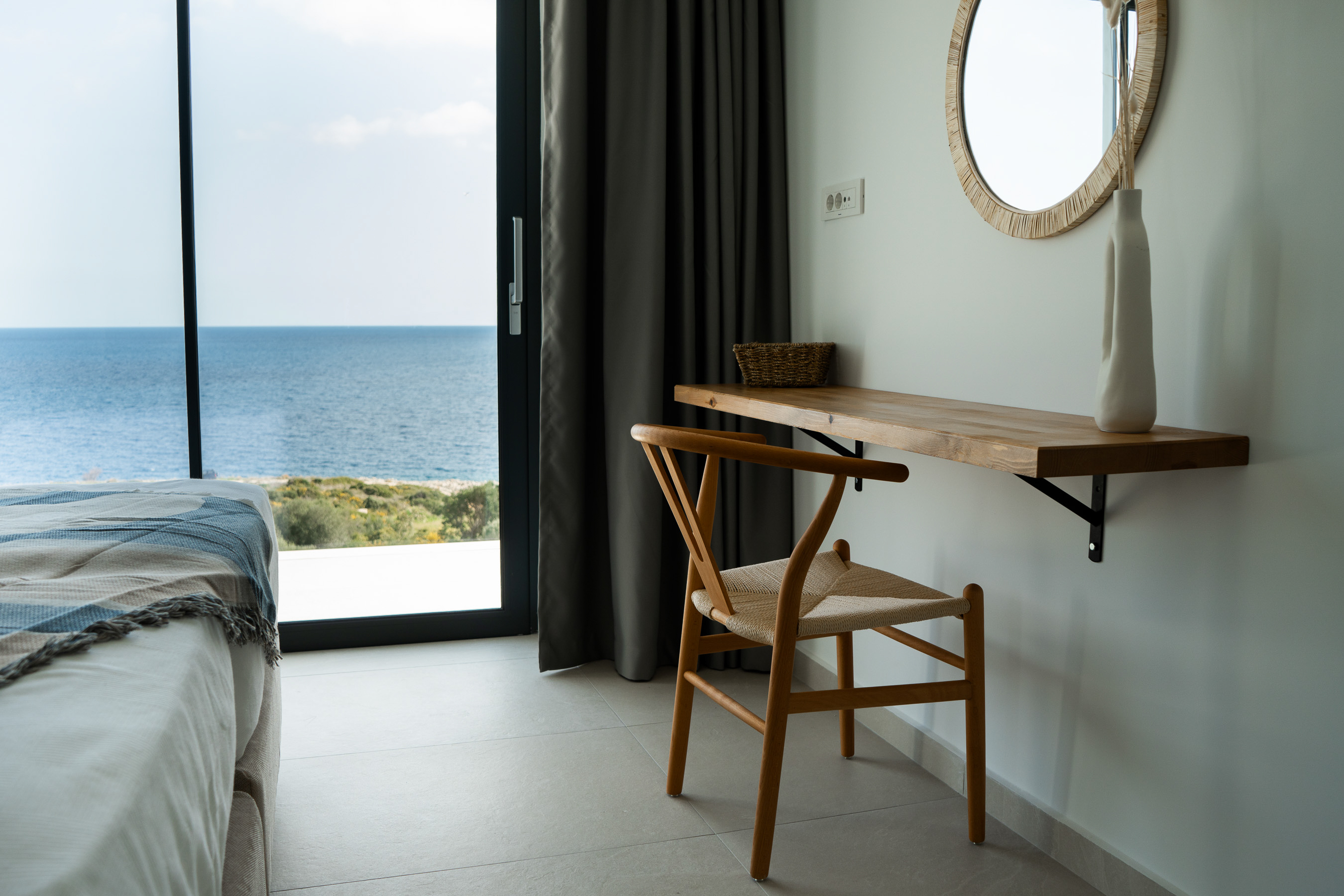 A bedroom study area features a wall-mounted timber desk with wishbone chair and round woven mirror. Large windows frame ocean views whilst maintaining the home's connection to the coastal setting.