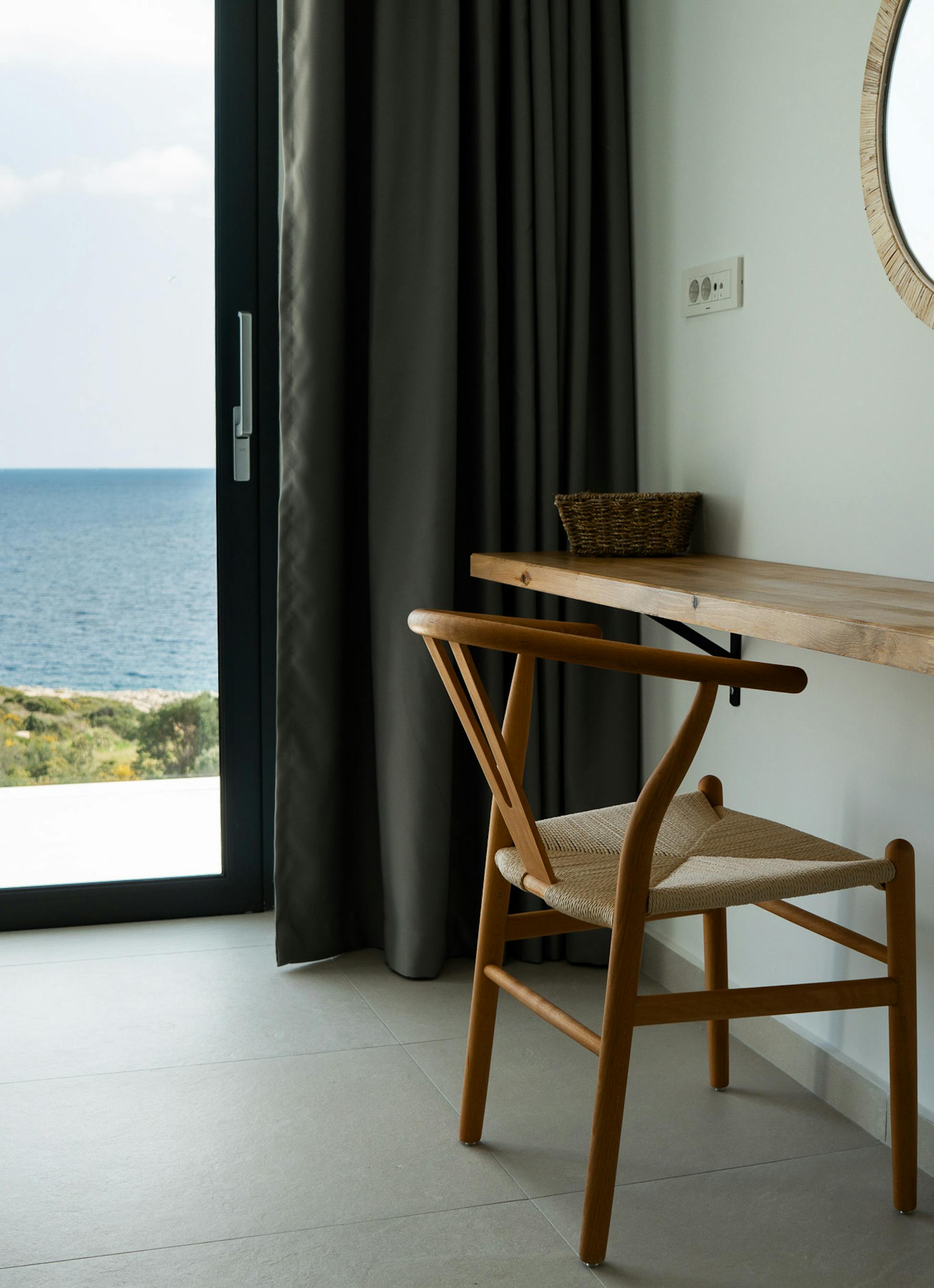 A bedroom study area features a wall-mounted timber desk with wishbone chair and round woven mirror. Large windows frame ocean views whilst maintaining the home's connection to the coastal setting.