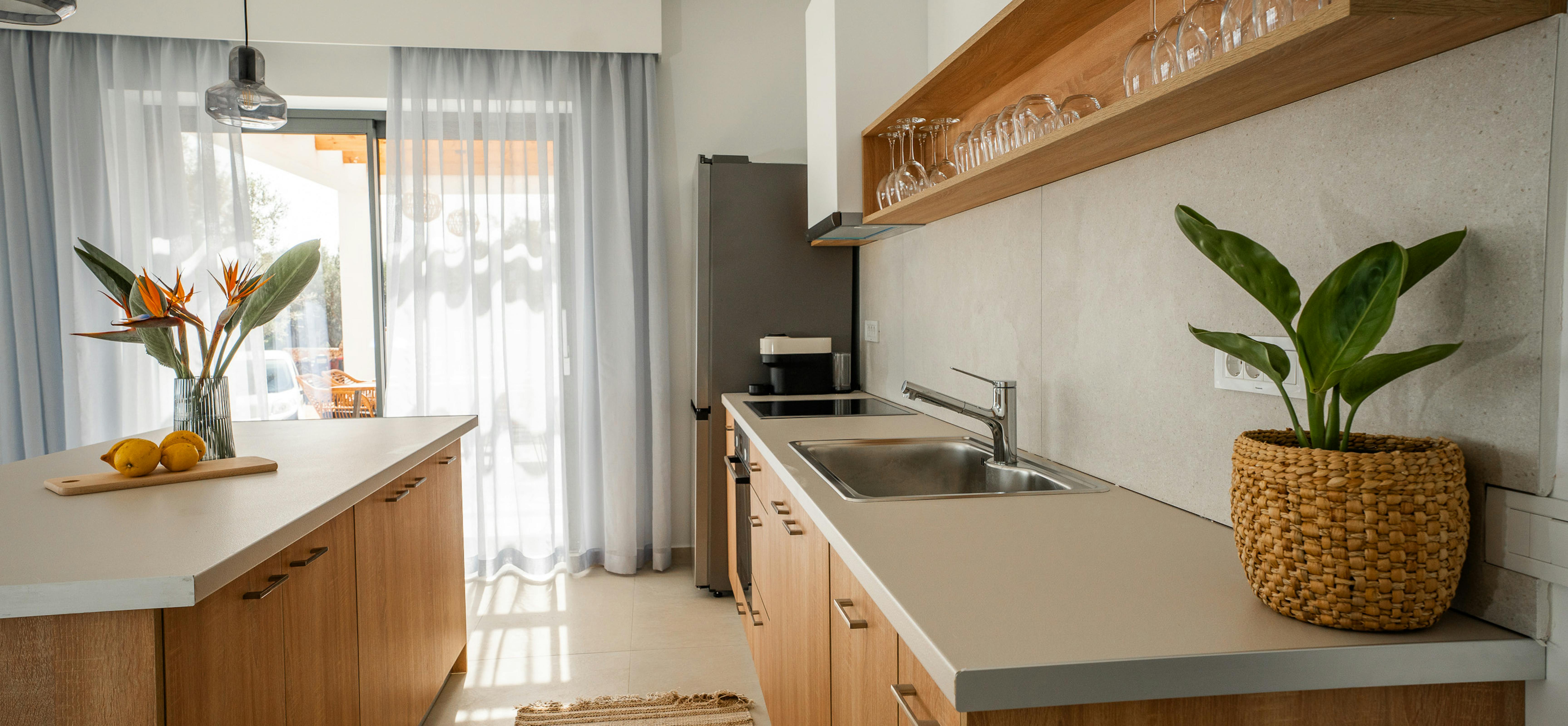 A modern galley kitchen features warm timber cabinetry with white stone bench tops and integrated appliances. Open timber shelving displays glassware whilst tropical plants and natural textures soften the contemporary design.