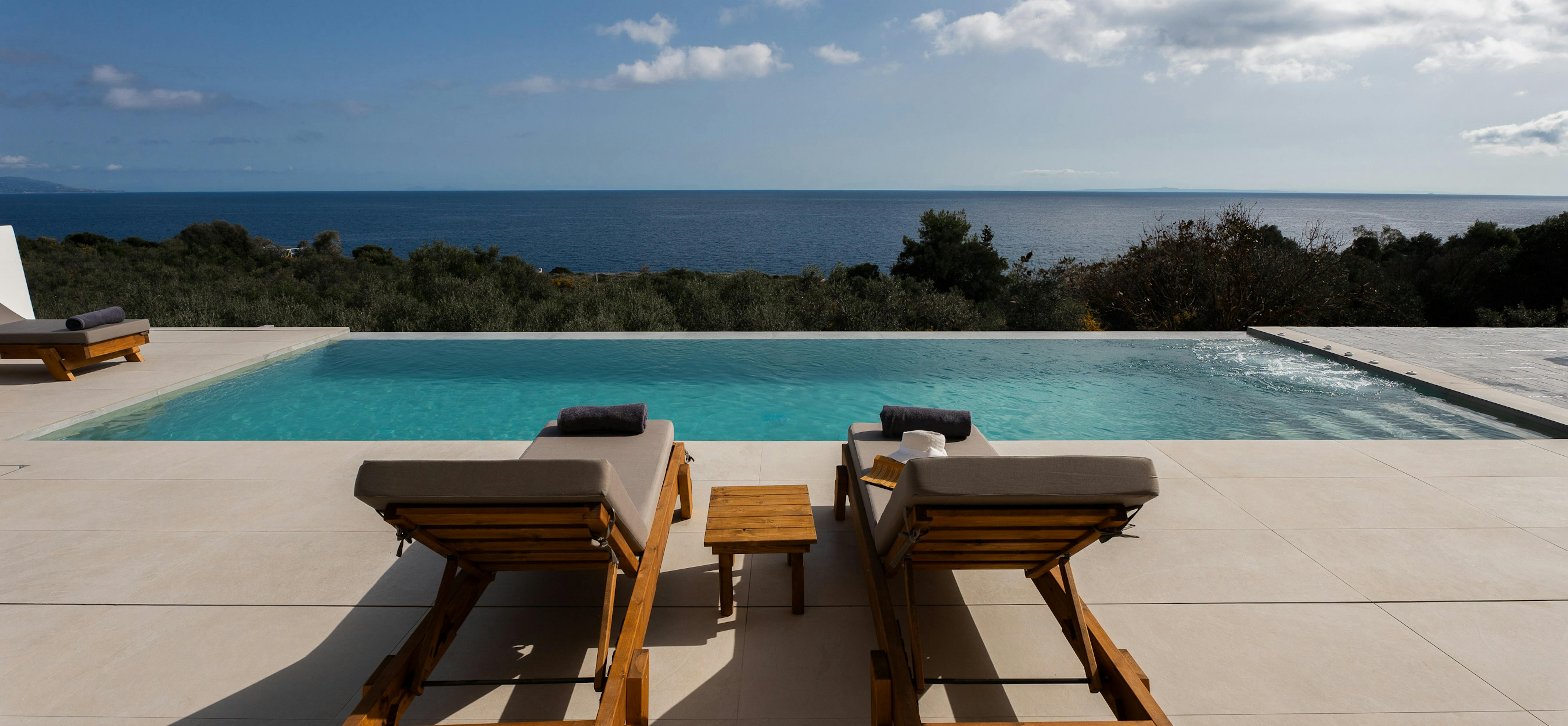 Two timber sun loungers are perfectly positioned beside an infinity pool with endless sea views. The symmetrical arrangement creates an inviting relaxation zone on the pristine white terrace.