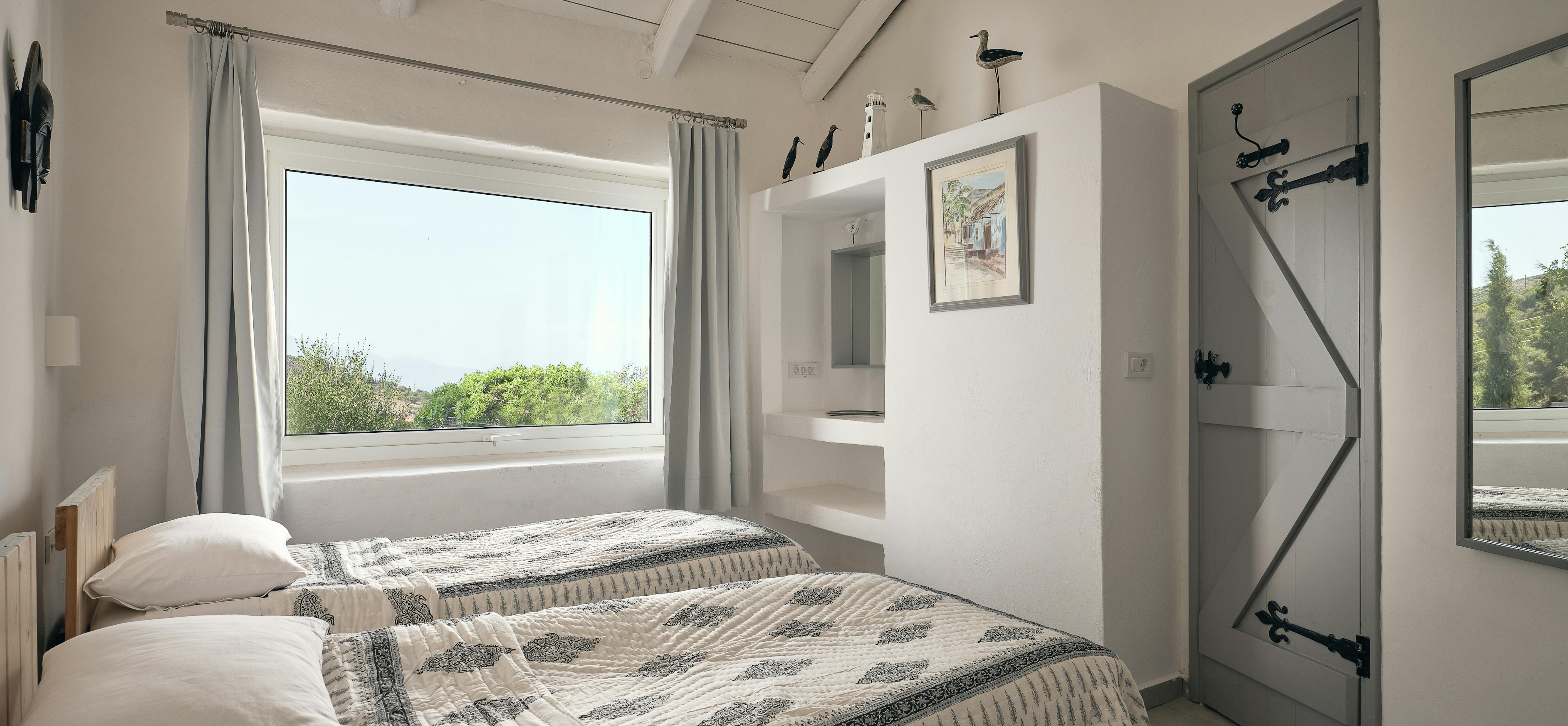 Twin beds with matching patterned throws are positioned beneath white-painted ceiling beams in a charming guest room. The nautical décor includes model boats, lighthouse ornaments, and coastal artwork creating seaside ambience.