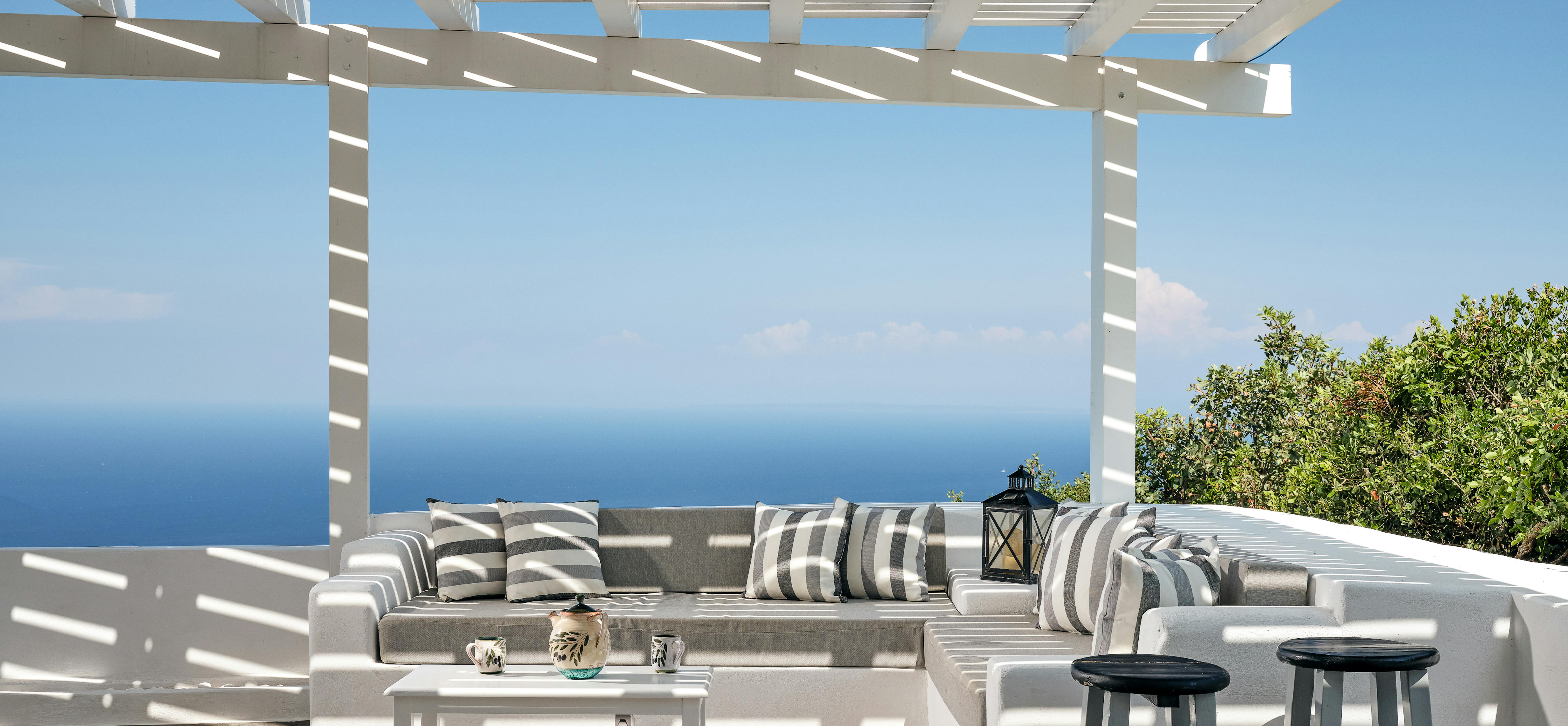 A covered terrace features white built-in seating with striped cushions overlooking spectacular Mediterranean sea views. The pergola structure provides shade whilst framing the endless blue horizon and coastal landscape below.