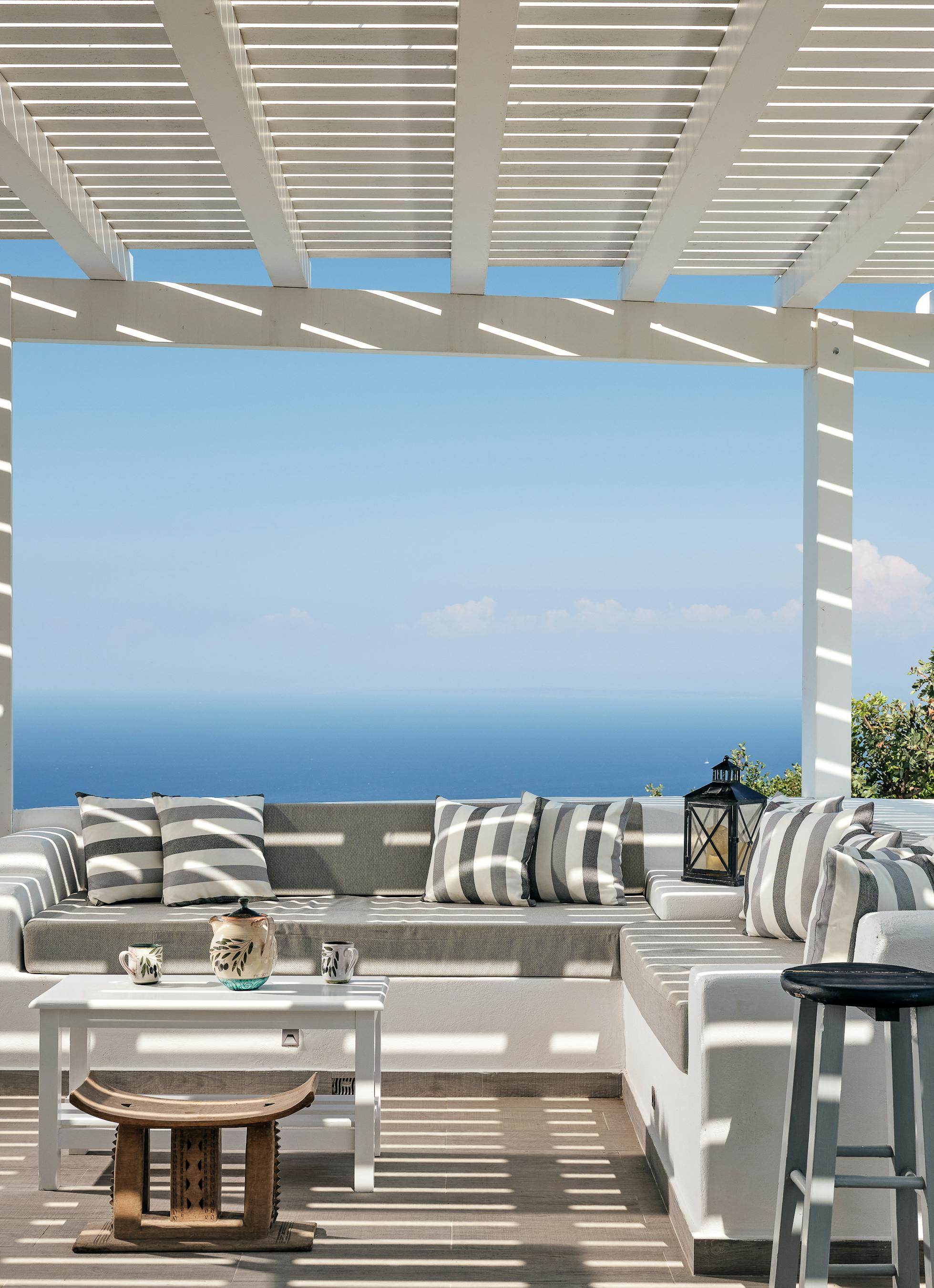 A covered terrace features white built-in seating with striped cushions overlooking spectacular Mediterranean sea views. The pergola structure provides shade whilst framing the endless blue horizon and coastal landscape below.