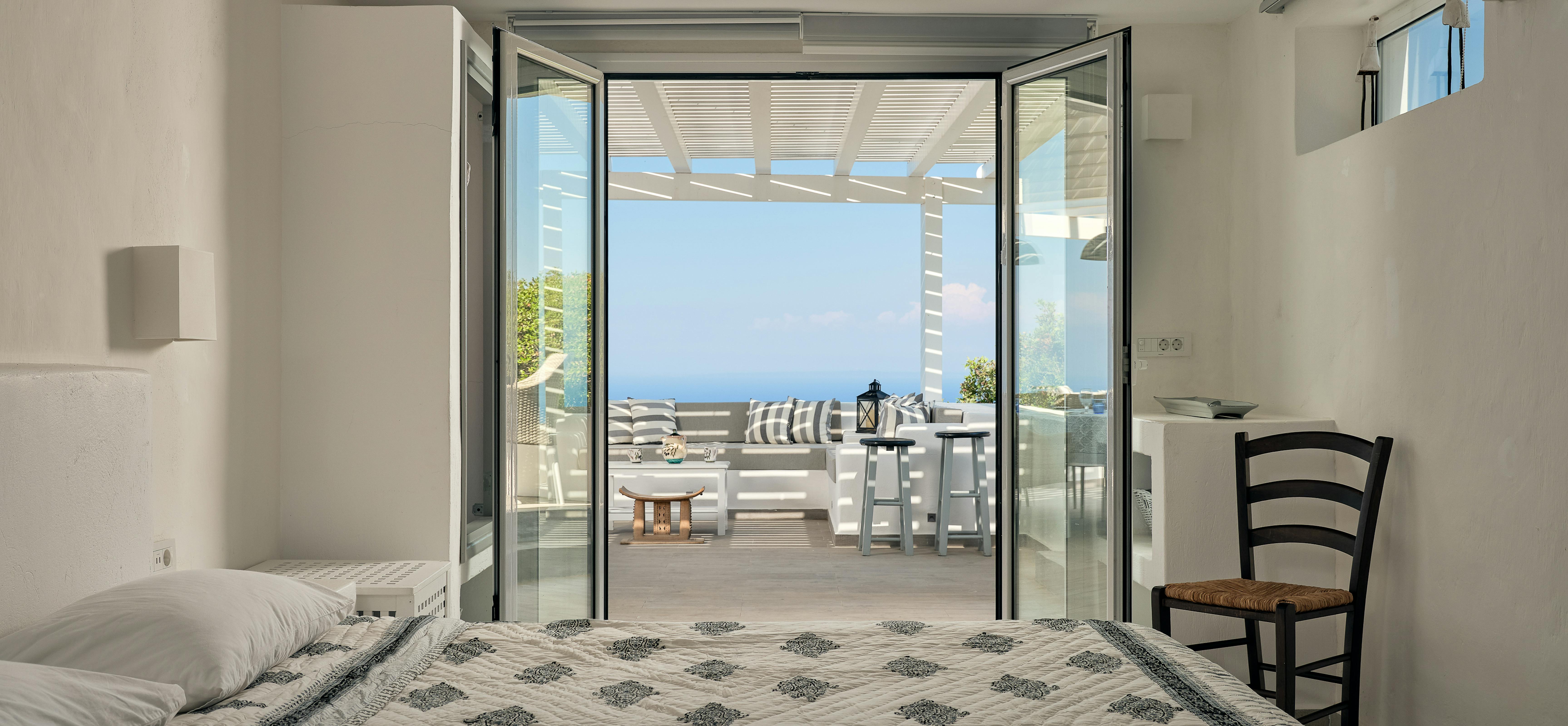 Floor-to-ceiling doors open from the bedroom to reveal a covered terrace with sea views and outdoor seating. The indoor-outdoor connection is enhanced by matching patterned textiles and neutral colour schemes throughout.