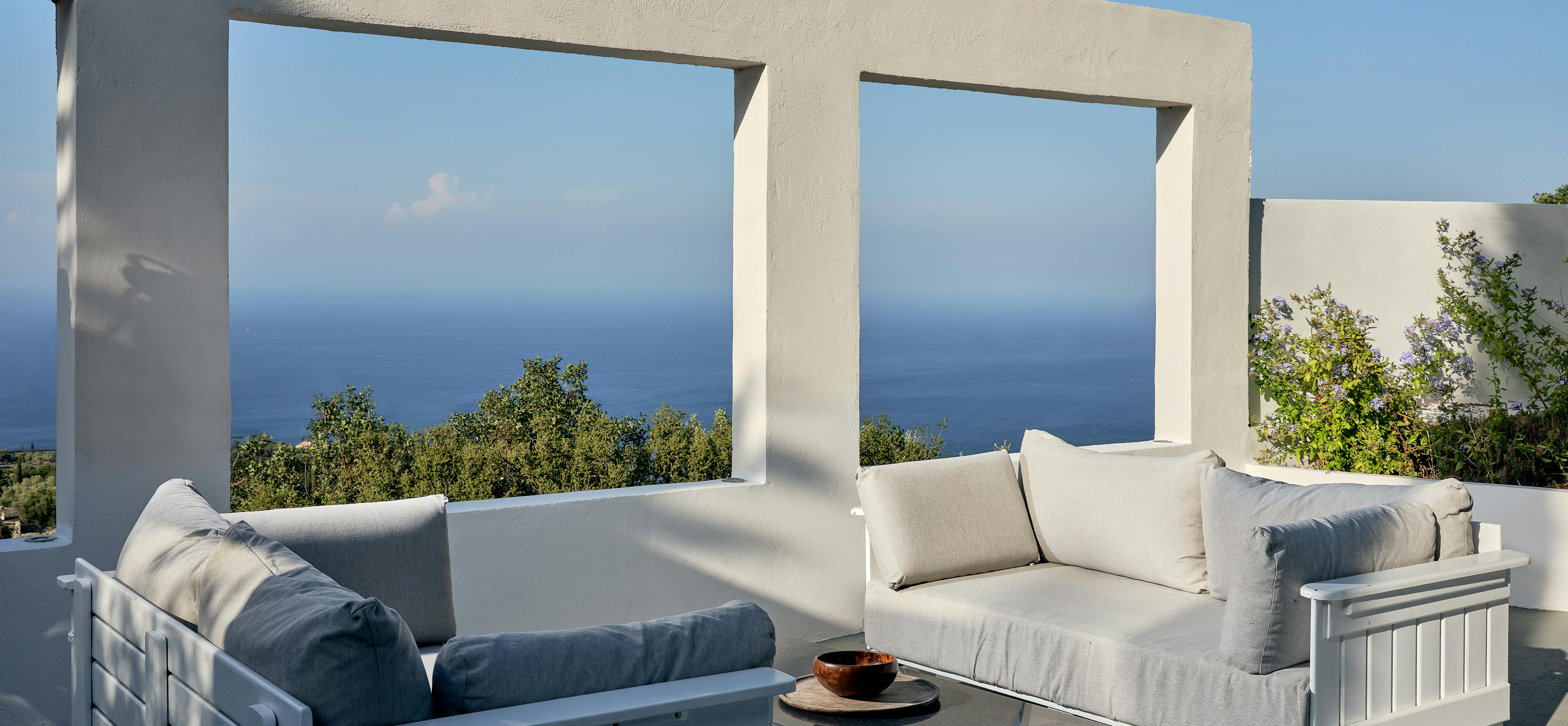 White timber outdoor furniture creates a sophisticated seating area on a rooftop terrace with panoramic sea views. The covered space provides shelter whilst maintaining connection to the spectacular Mediterranean coastline beyond.