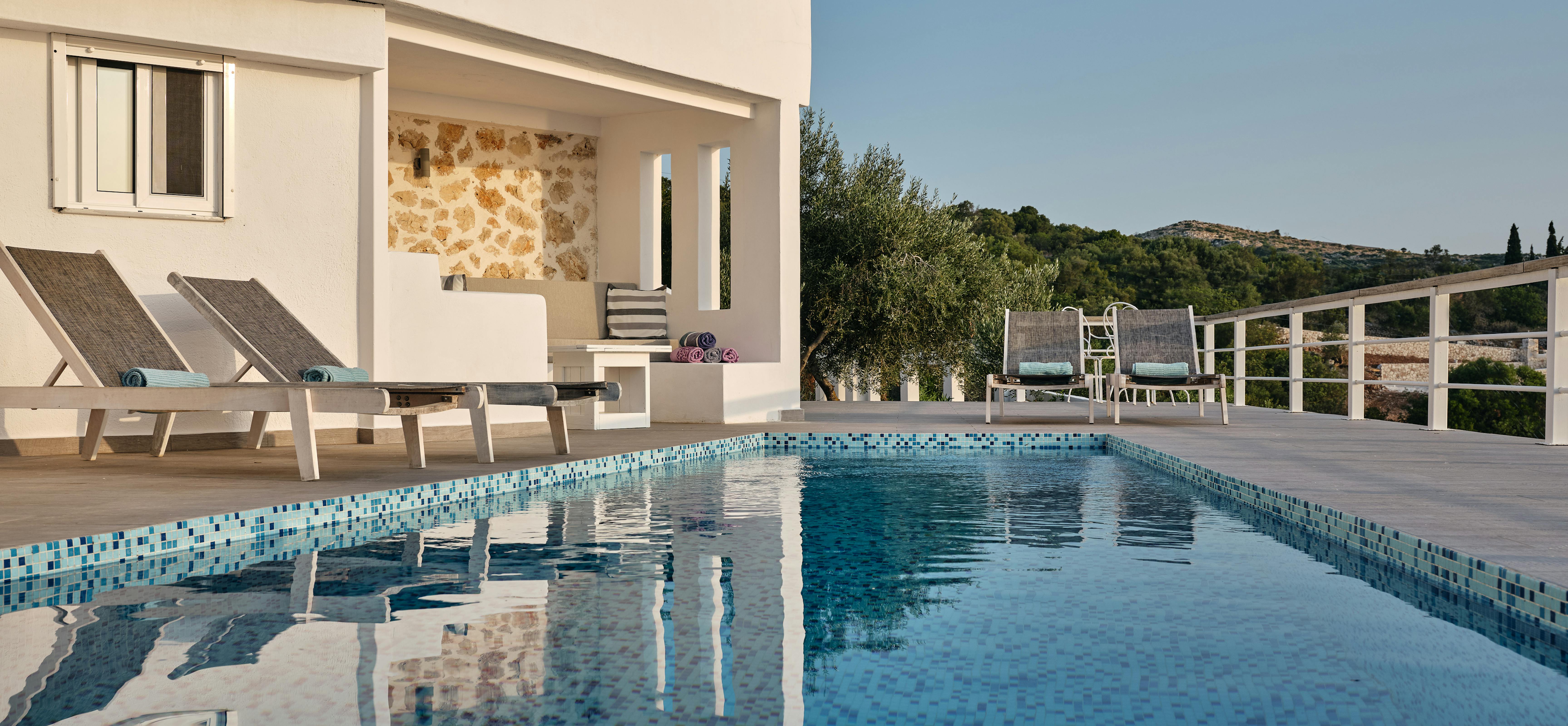 A modern Mediterranean villa features a blue mosaic-tiled swimming pool reflecting the white architecture and hillside setting. White sun loungers are positioned around the pool terrace with natural stone accents and covered outdoor living areas.