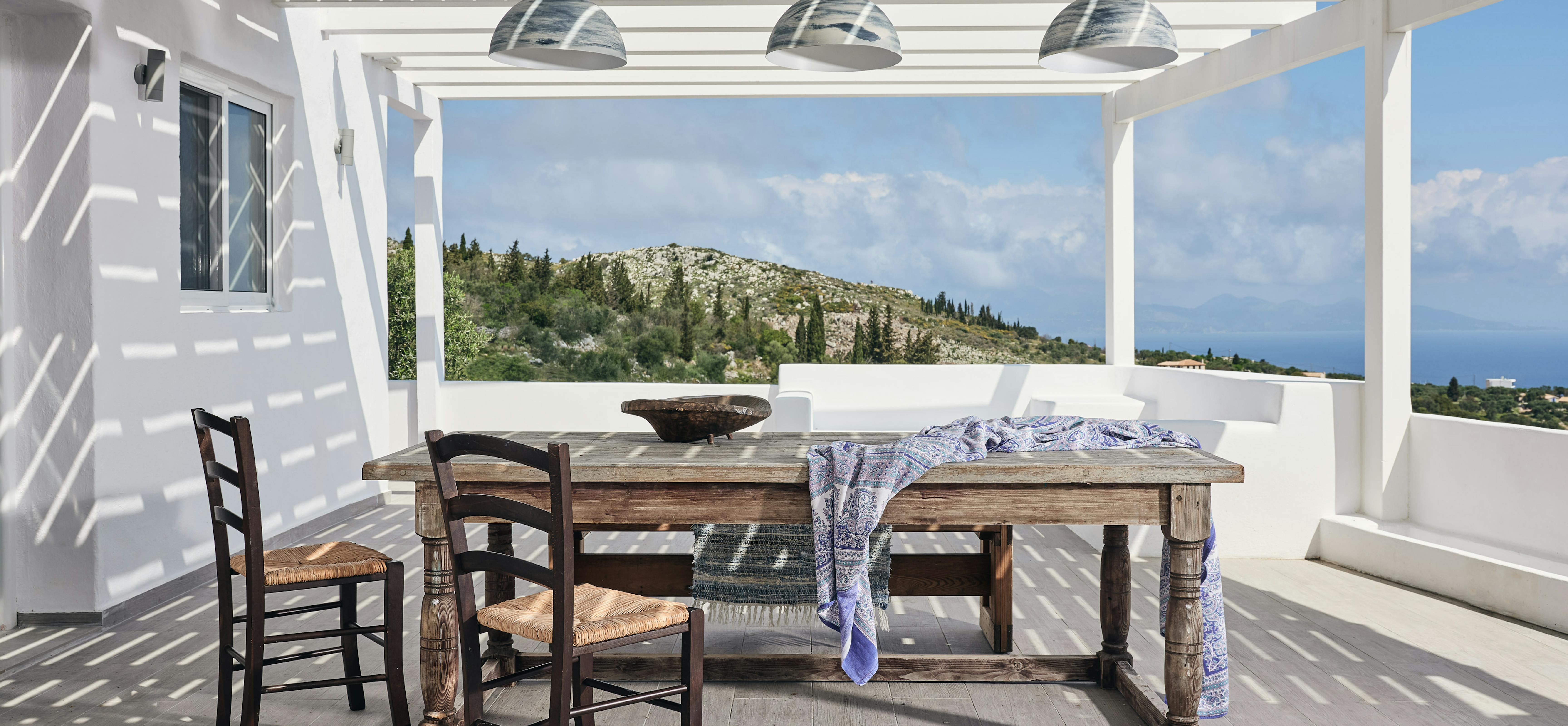 Traditional rush-seated chairs surround a rustic timber dining table on a covered terrace with industrial pendant lighting. The white pergola structure frames views of the Mediterranean landscape and creates dramatic shadow patterns across the deck.