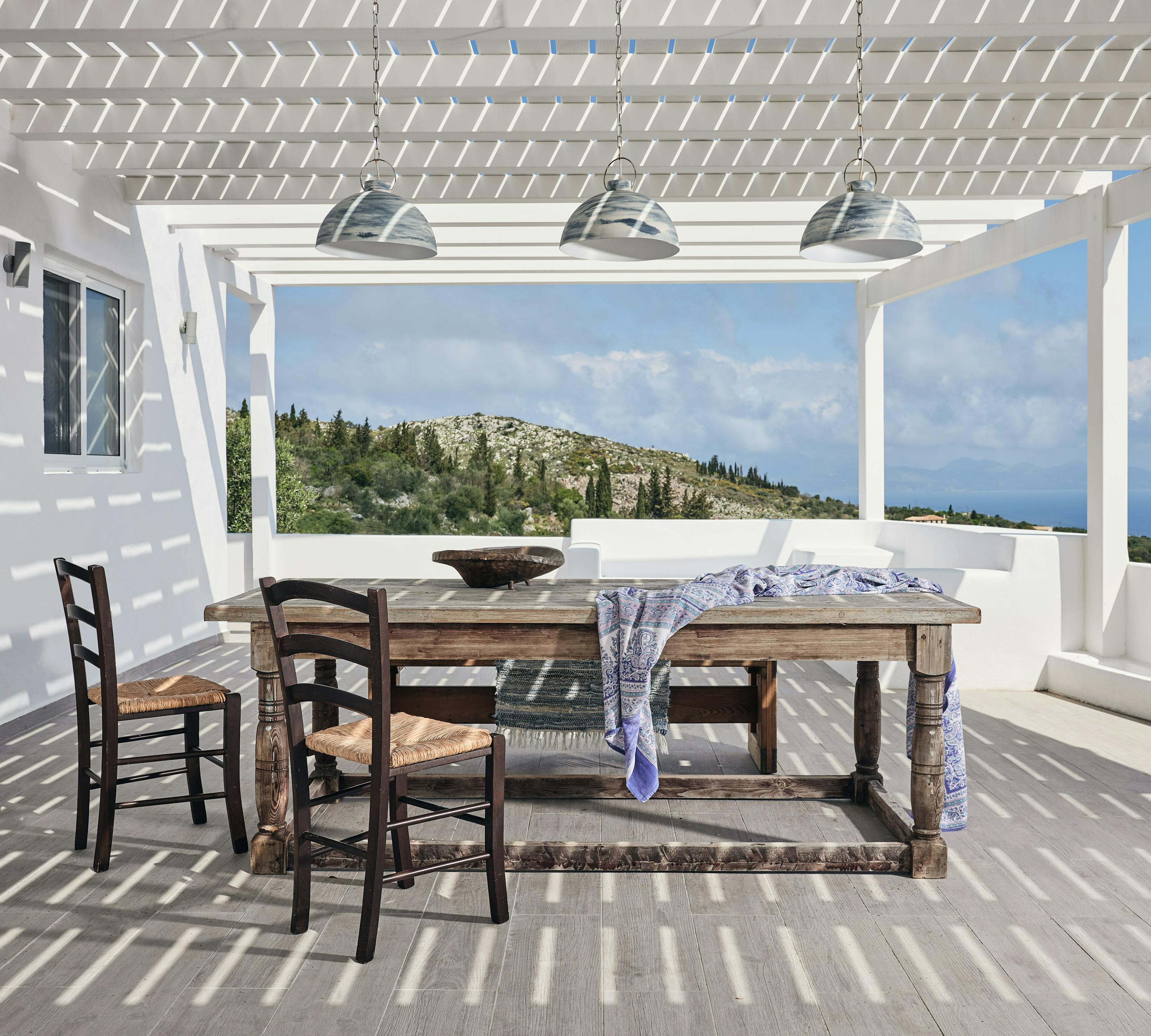 Traditional rush-seated chairs surround a rustic timber dining table on a covered terrace with industrial pendant lighting. The white pergola structure frames views of the Mediterranean landscape and creates dramatic shadow patterns across the deck.