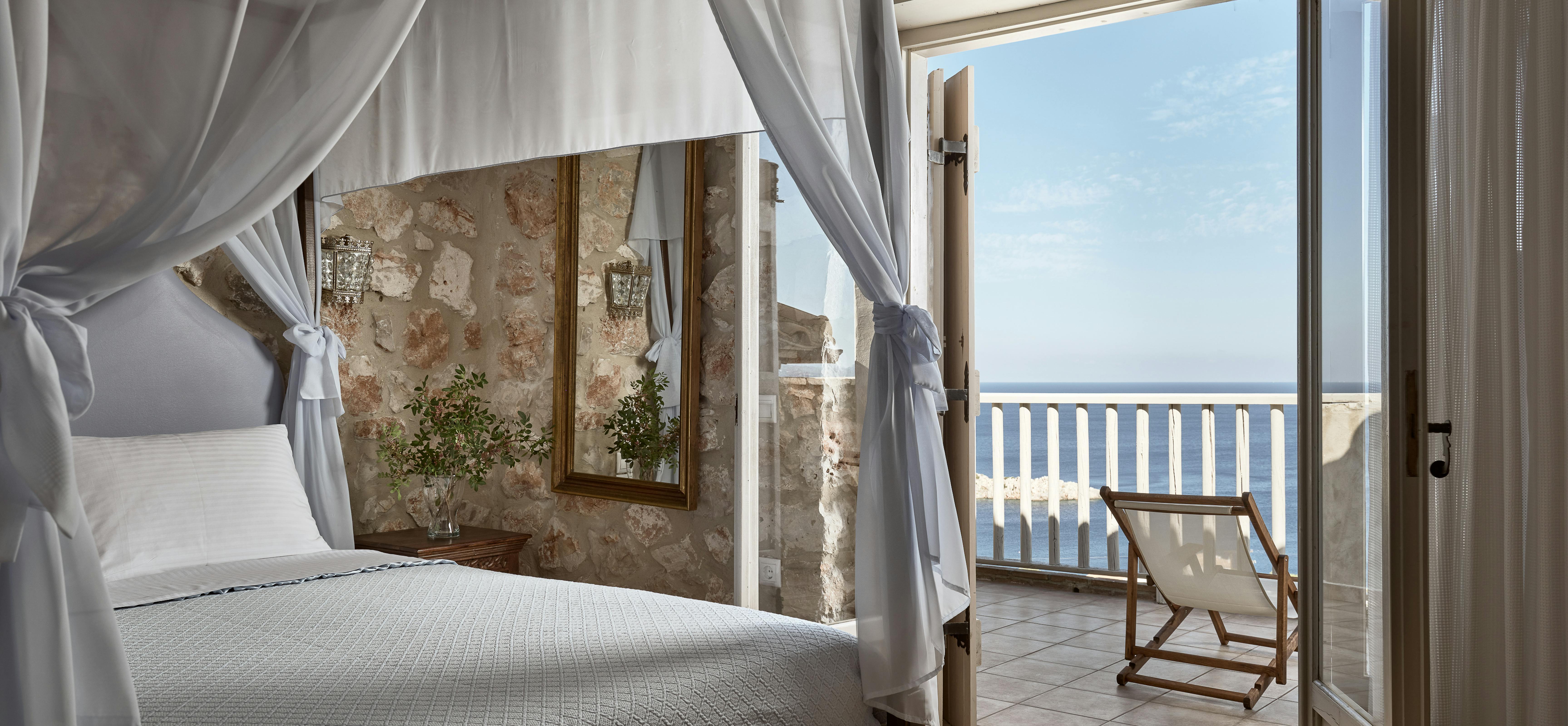 White curtains drape around the timber four-poster bed frame, creating an intimate sleeping sanctuary within the stone-walled room. French doors provide access to a private balcony overlooking the endless blue Mediterranean horizon.