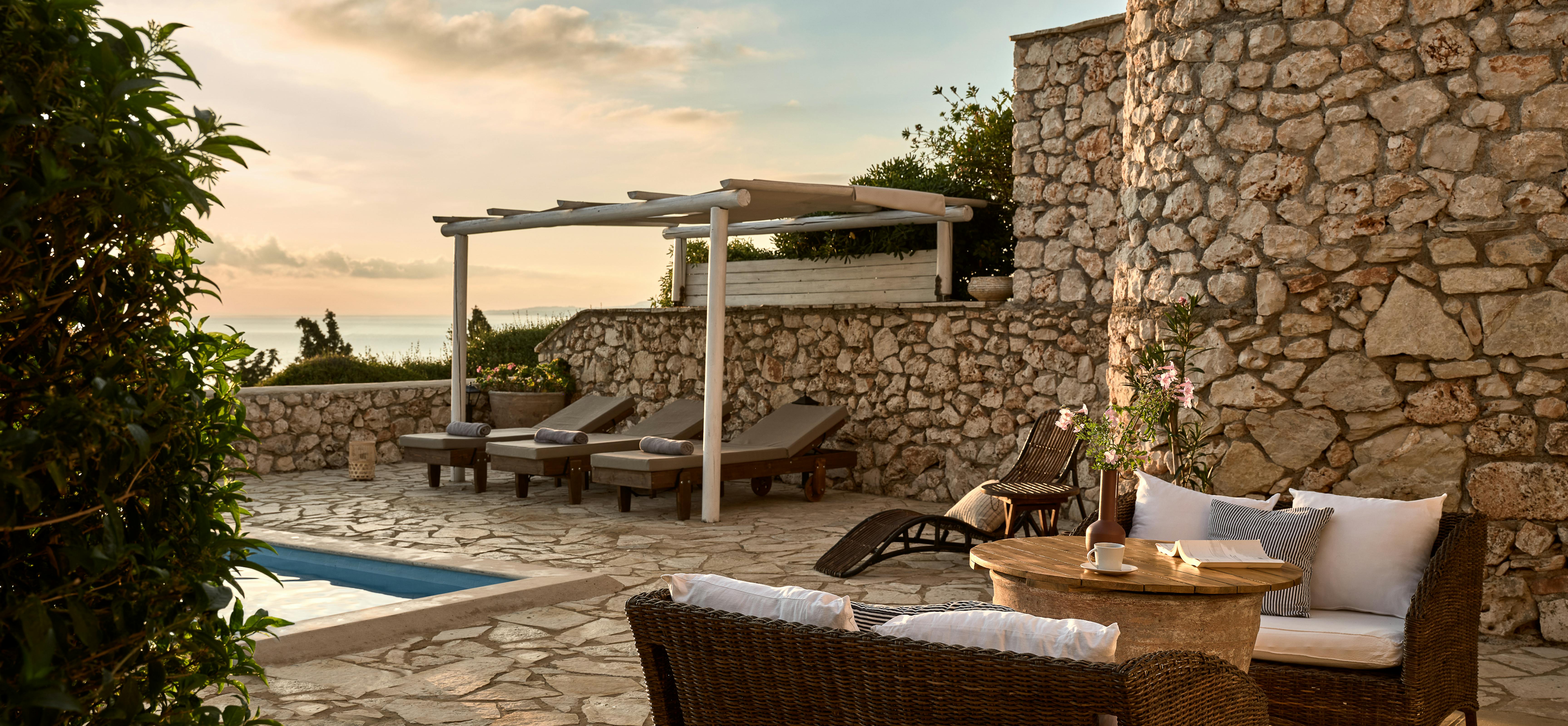 A stone-paved terrace features wicker outdoor furniture positioned to capture golden hour light and sea views. Natural stone walls provide shelter whilst the infinity pool reflects the surrounding Mediterranean vegetation and coastline.