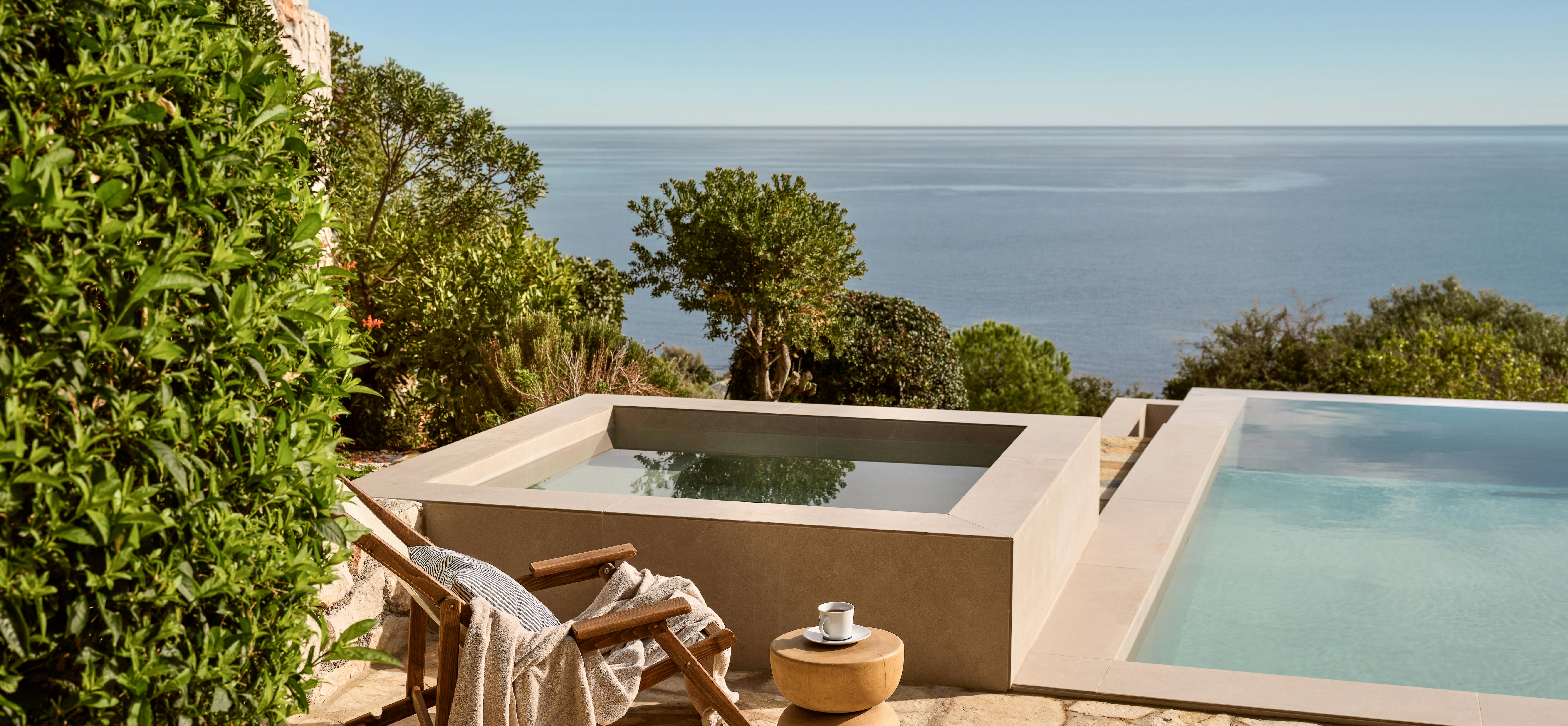 Timber sun loungers overlook both an infinity pool and the Mediterranean coastline from this elevated coastal position. A sculptural side table complements the natural stone terrace materials and lush Mediterranean vegetation.