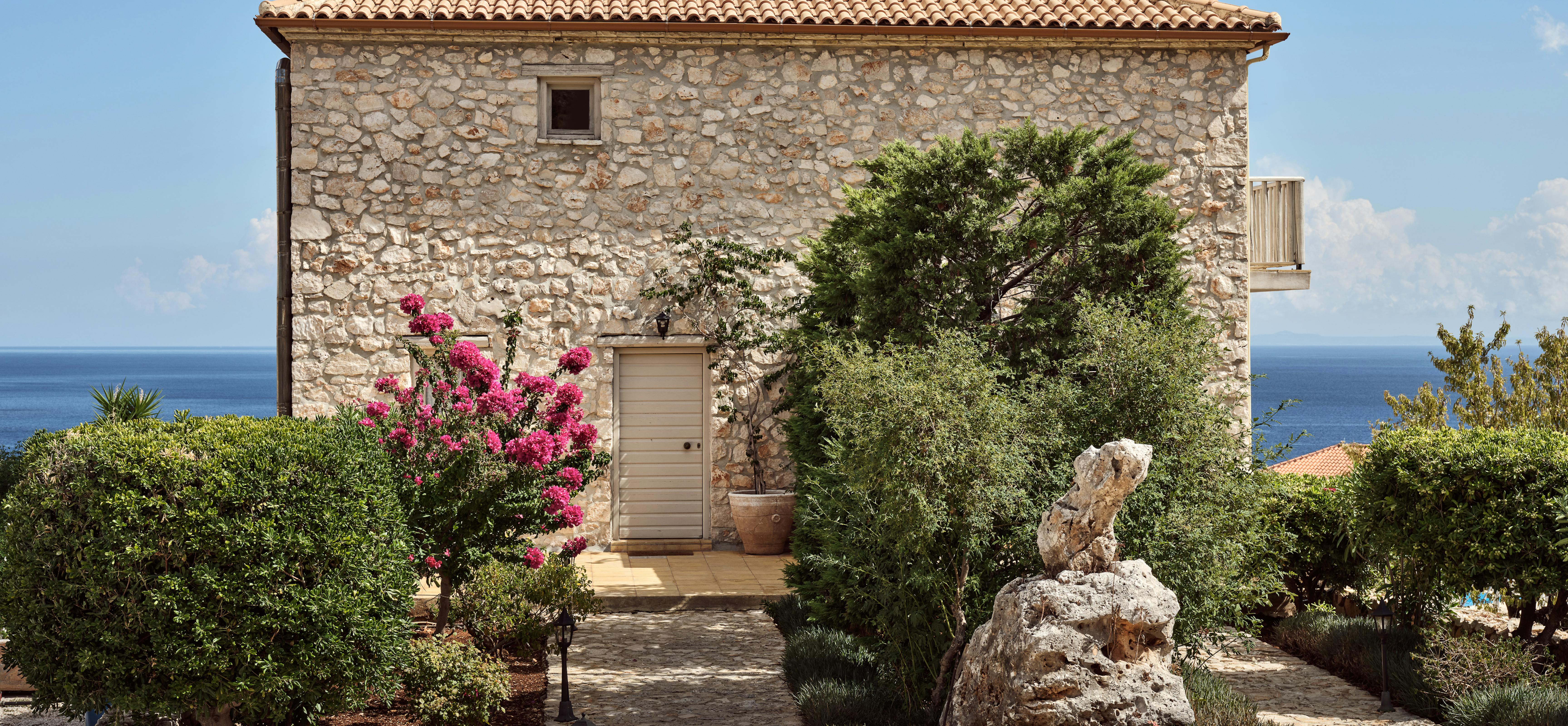 A traditional Mediterranean stone villa with terracotta roof tiles sits amongst landscaped gardens overlooking the sea. Vibrant pink bougainvillea and mature olive trees frame the cobblestone courtyard and rustic entrance.