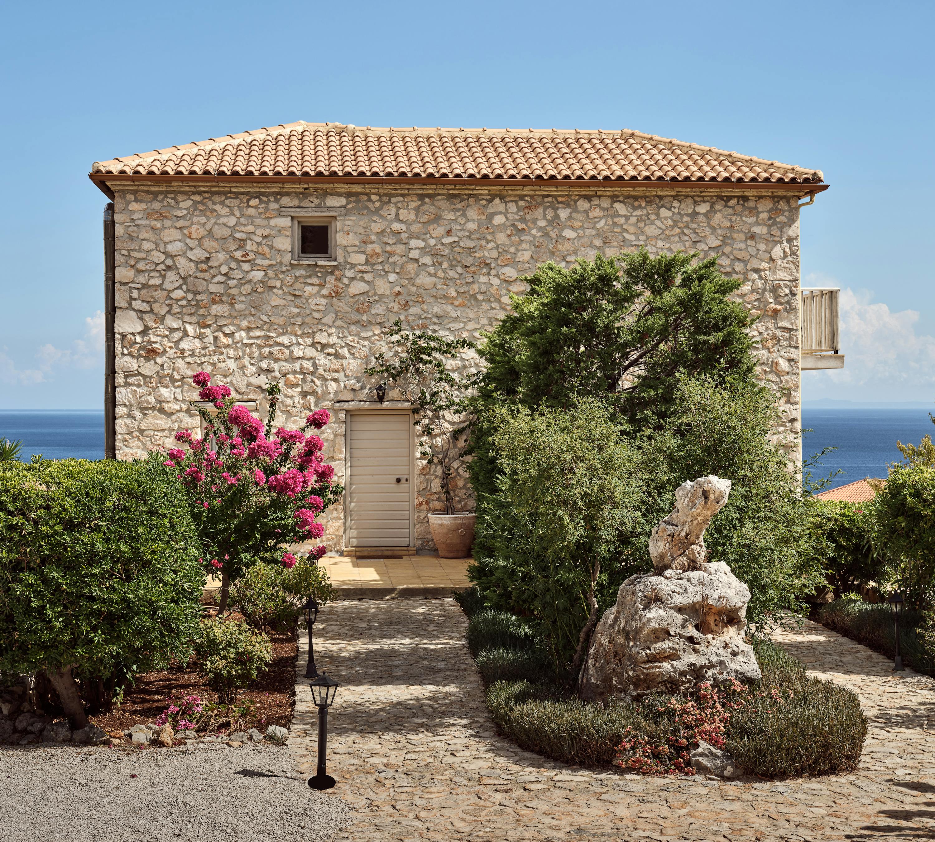 A traditional Mediterranean stone villa with terracotta roof tiles sits amongst landscaped gardens overlooking the sea. Vibrant pink bougainvillea and mature olive trees frame the cobblestone courtyard and rustic entrance.