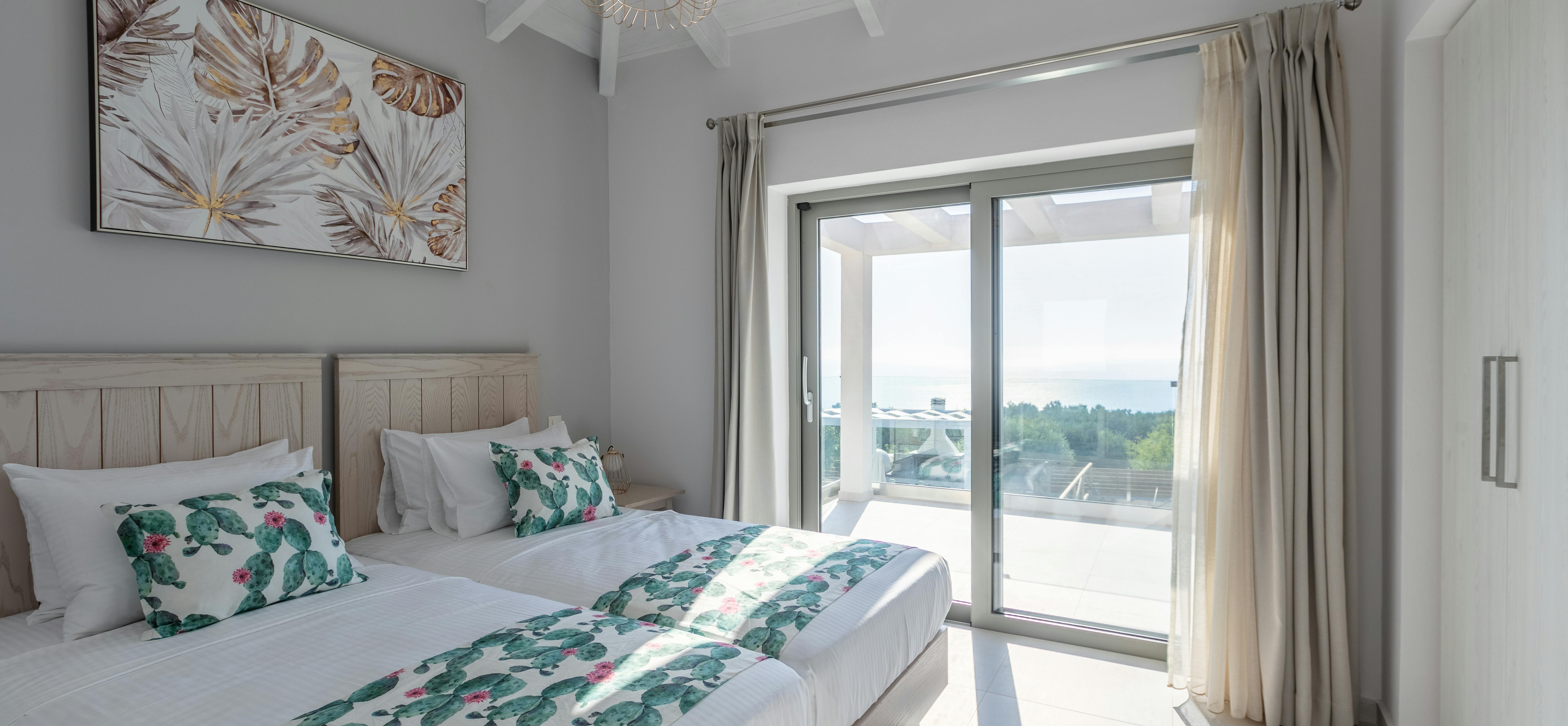 Bedroom with vaulted ceiling, wicker pendant light, tropical leaf artwork, bed with green floral bedding, white furnishings, and sliding doors opening to sunny terrace.