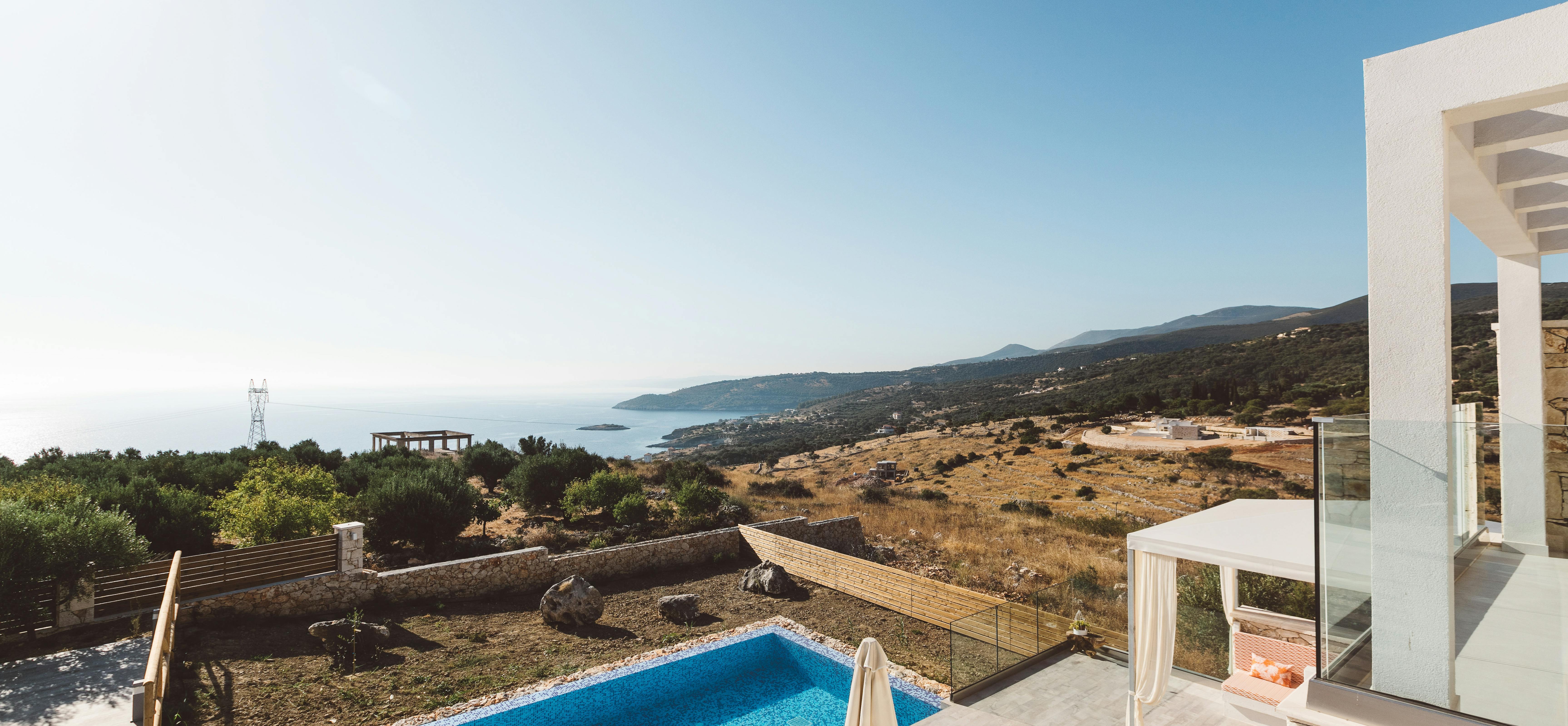 Elevated pool deck with glass railings, white umbrella, single lounger, and sweeping views of terraced hillsides descending toward sparkling turquoise bay.