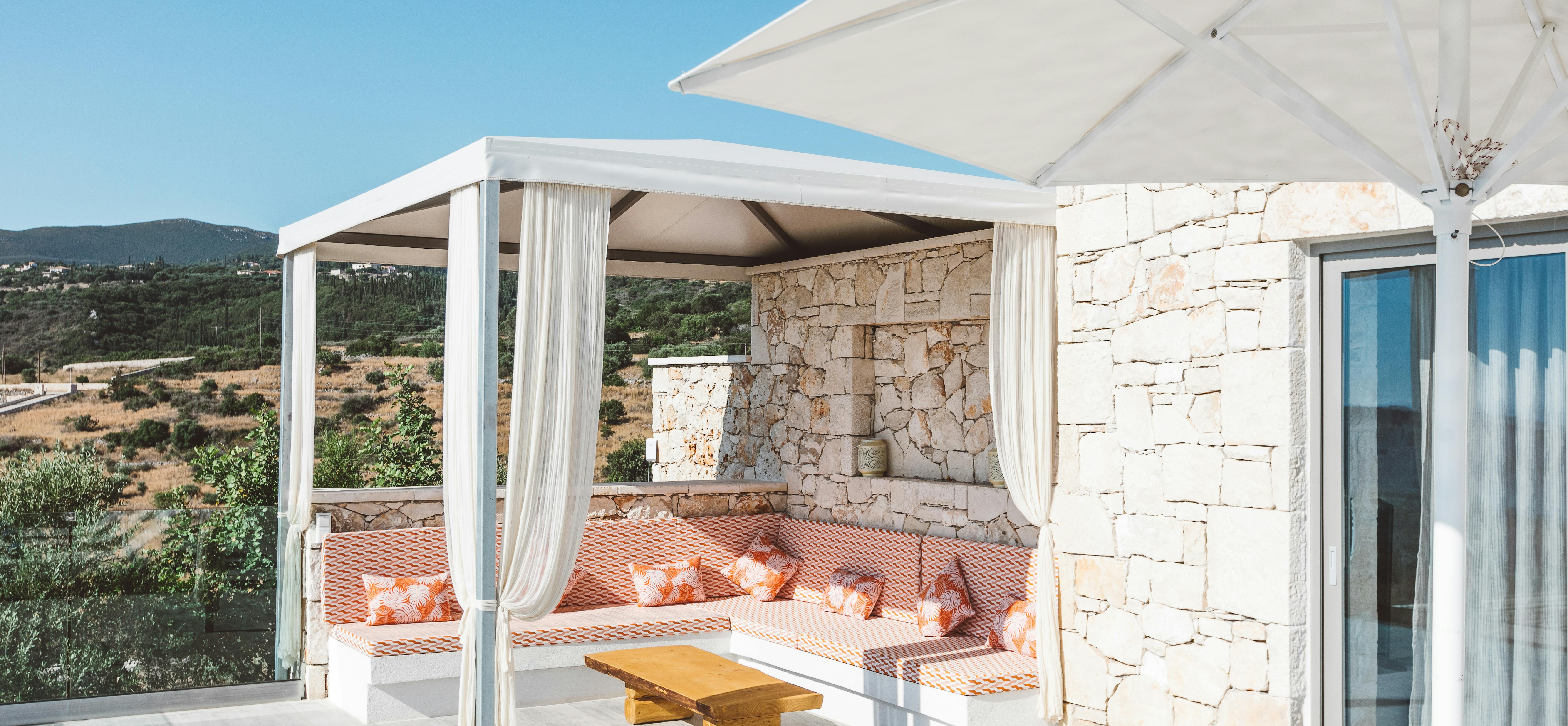 Shaded lounge pavilion with white frame, flowing curtains, built-in orange-cushioned seating, wooden coffee table, and open views of coastal landscape beyond stone walls.