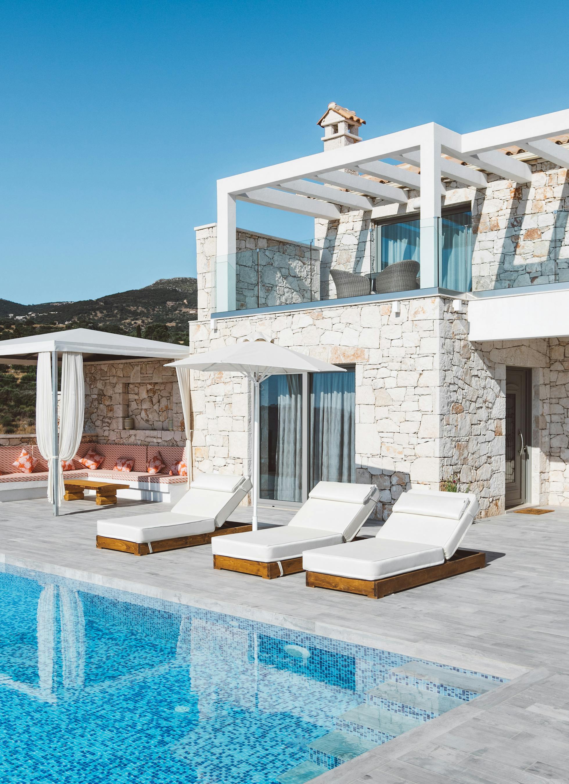 Two-story stone villa with white pergolas, infinity pool with submerged steps, white loungers, draped daybed pavilion, and mountain backdrop under clear blue sky.