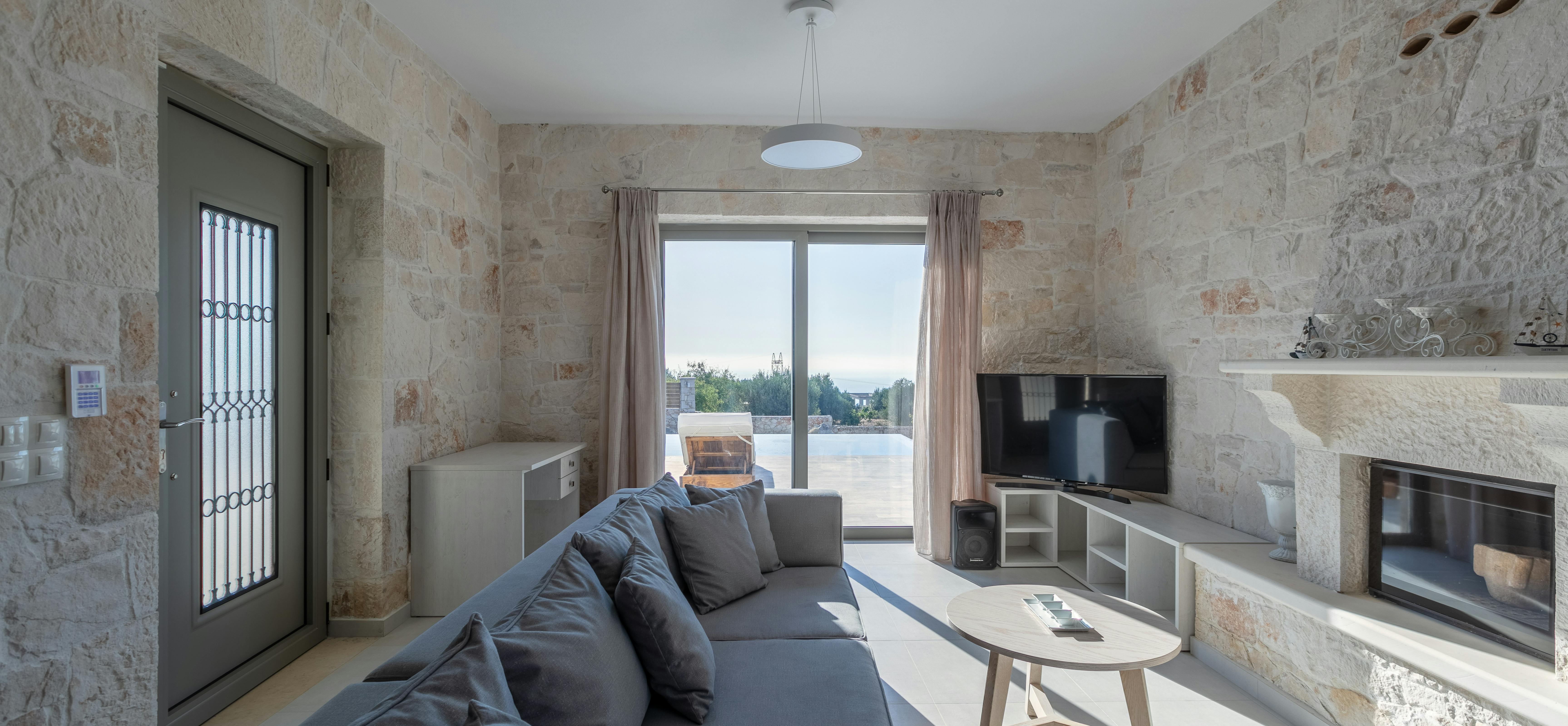 Rustic living room with exposed stone walls, grey sectional sofa, oval coffee table, built-in fireplace, TV console, and bright terrace doors framing coastal vista.
