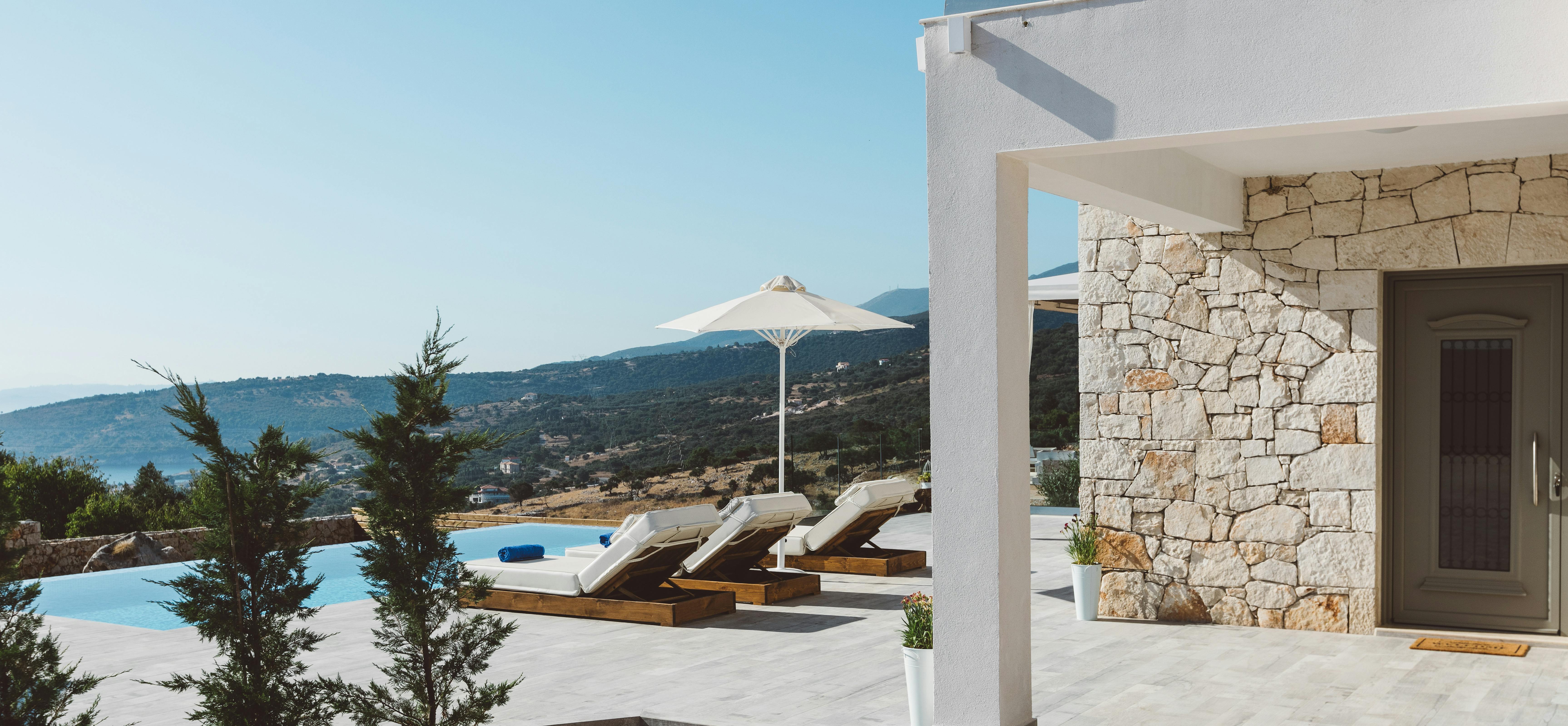Modern villa entrance with stone facade, white umbrella, wooden loungers by infinity pool, white pergola, cypress trees, and panoramic hillside and coastal views.