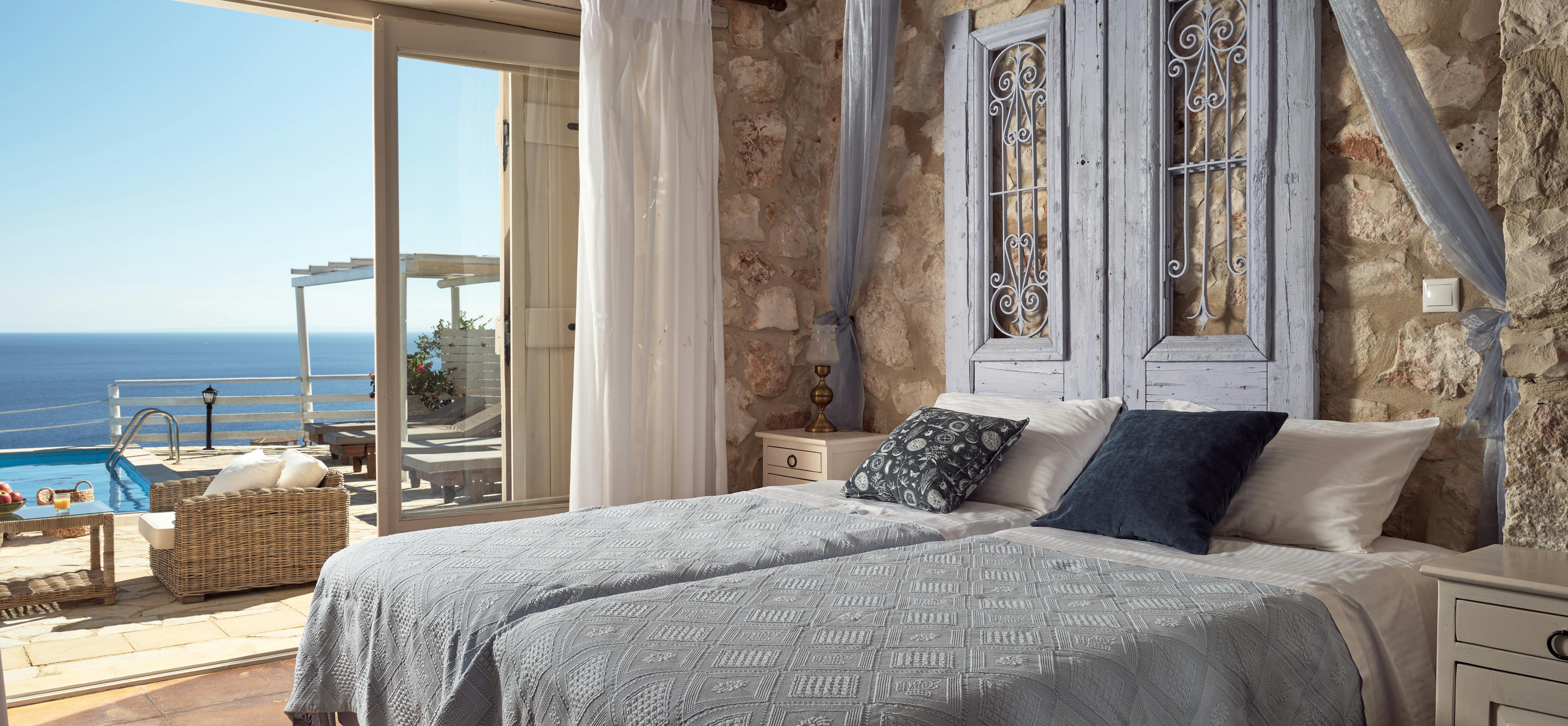 Rustic stone bedroom with ocean views, white canopy bed, and French doors opening to terrace with pool.