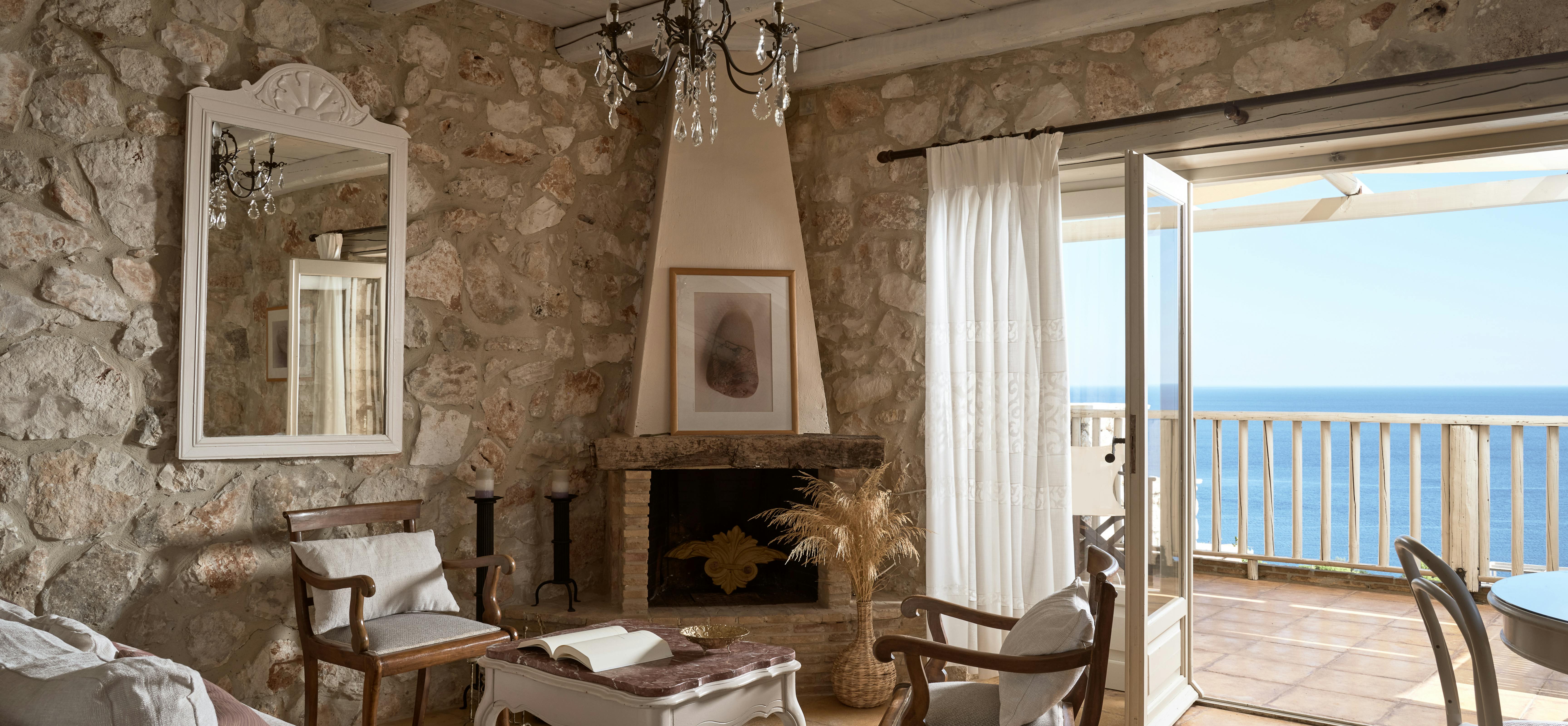 Living room with stone fireplace, vintage chandelier, and French doors opening to oceanfront balcony.
