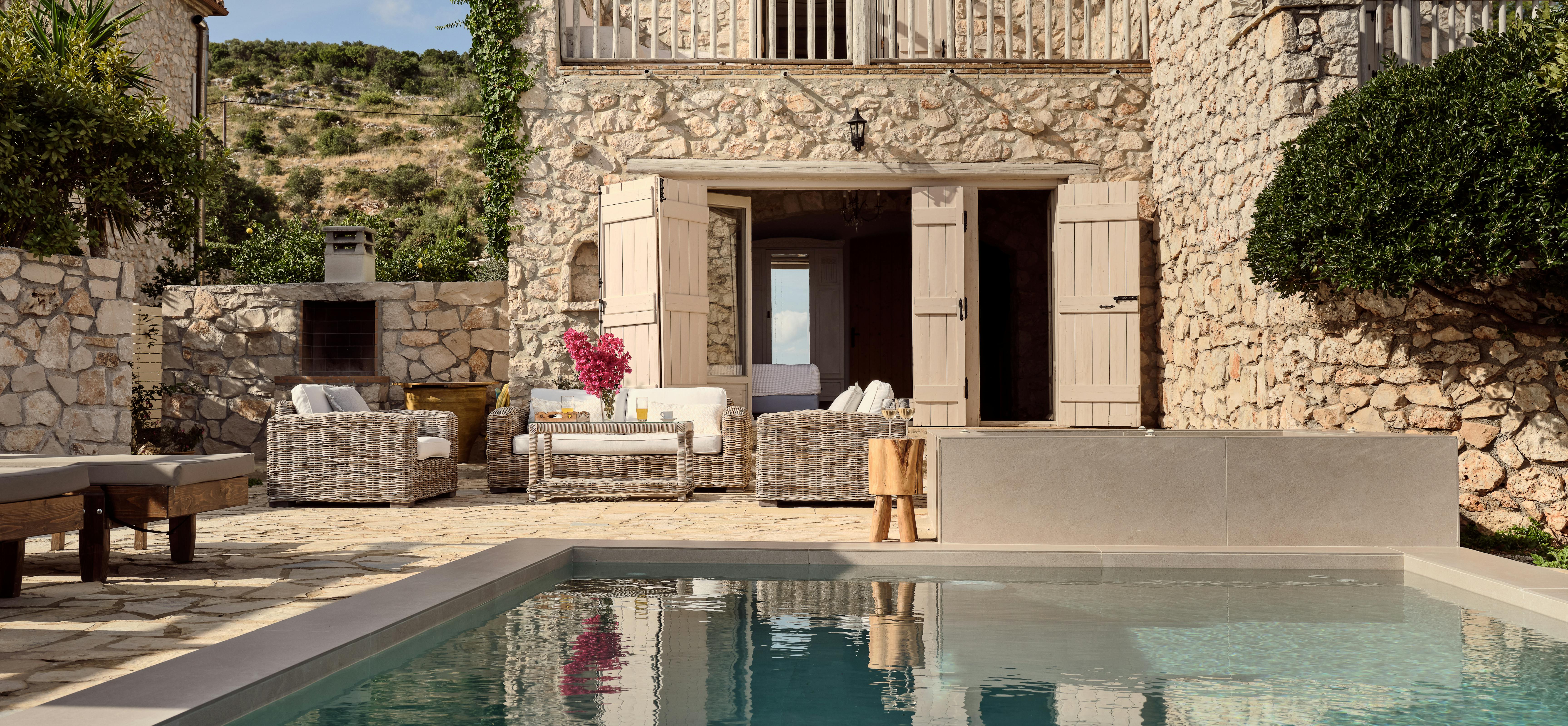 Two-story stone villa with upper balcony, pool terrace, and wicker outdoor furniture.