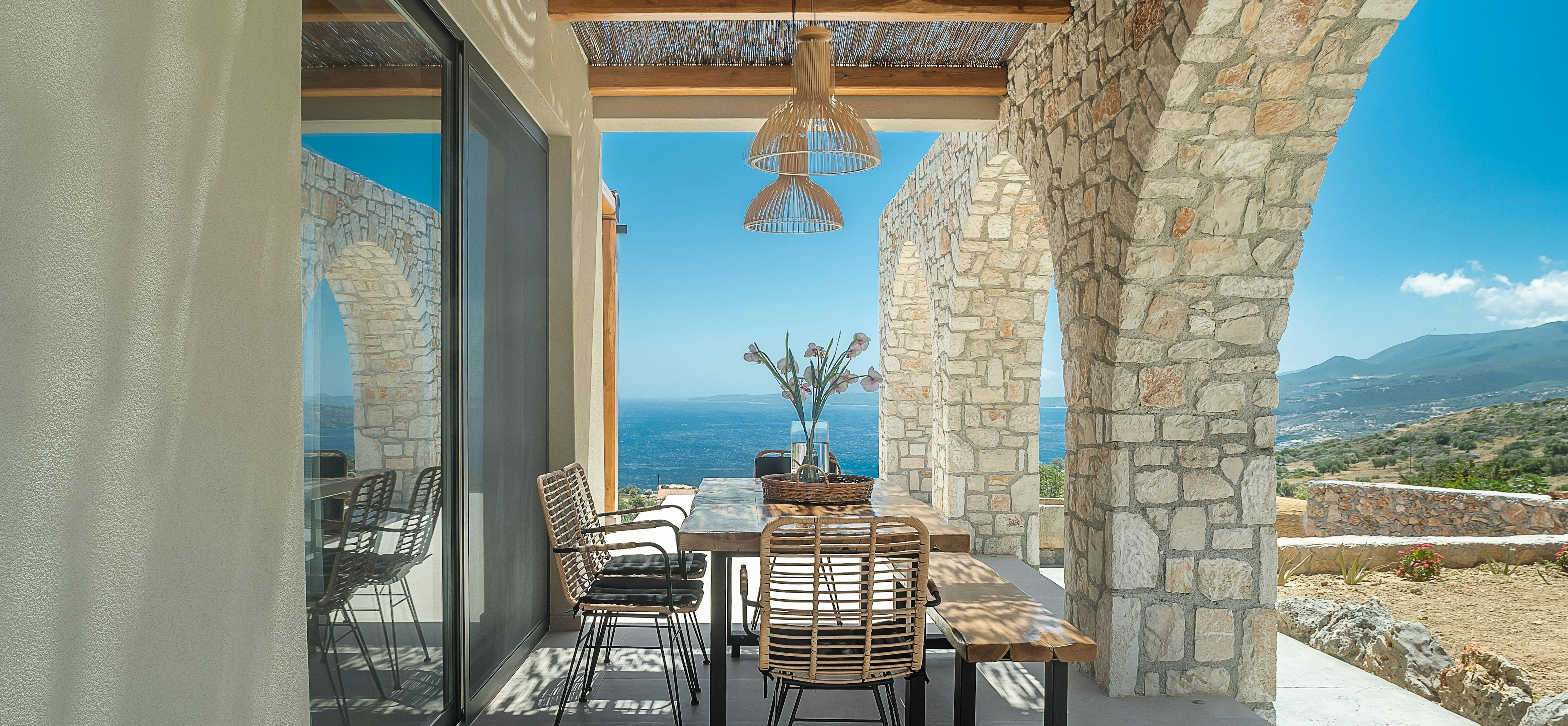 Outdoor dining area with wooden table under pergola, stone columns, and ocean vistas.