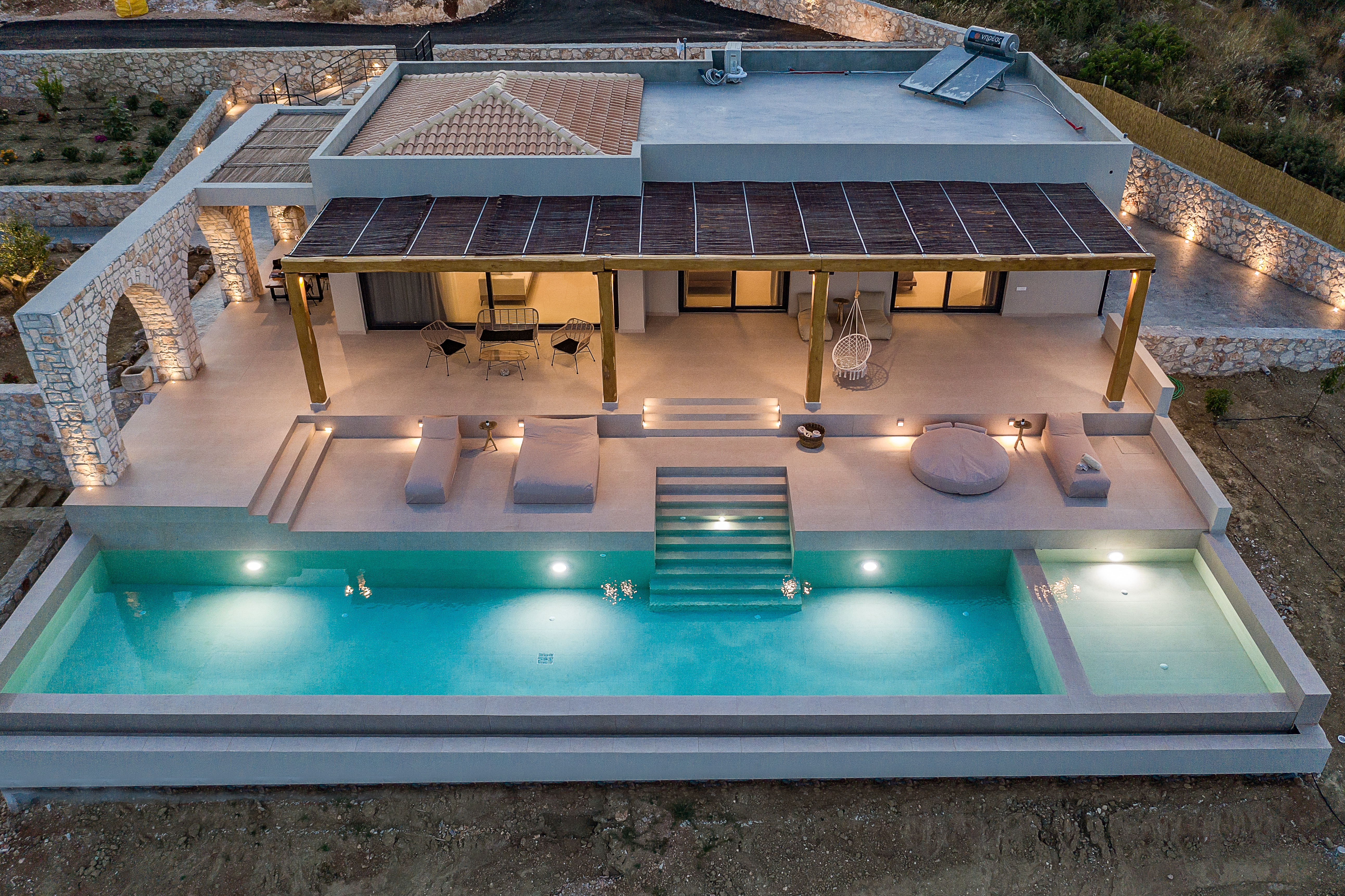 Aerial view of modern villa with rectangular infinity pool, illuminated terraces, and stone archways.