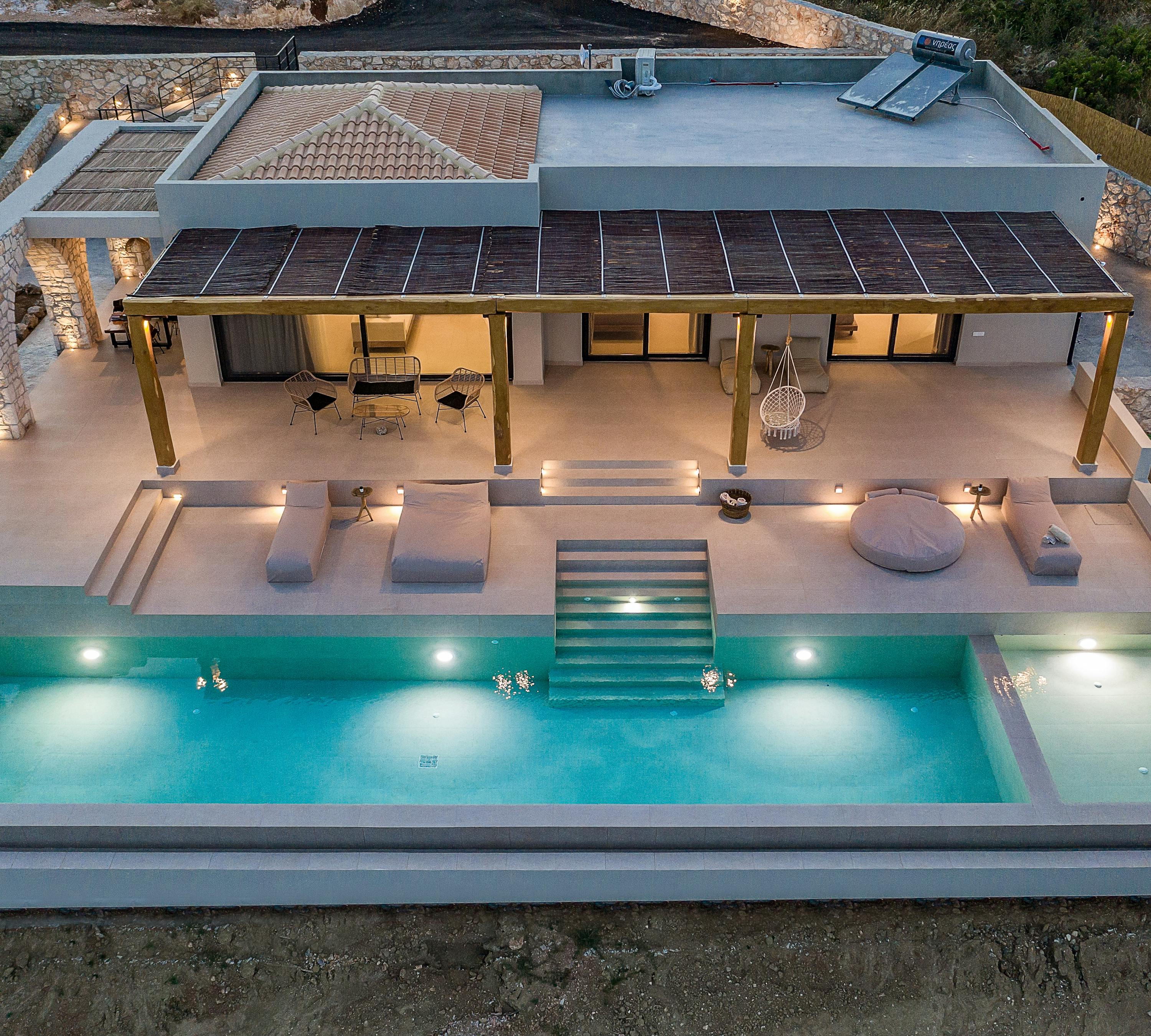Aerial view of modern villa with rectangular infinity pool, illuminated terraces, and stone archways.