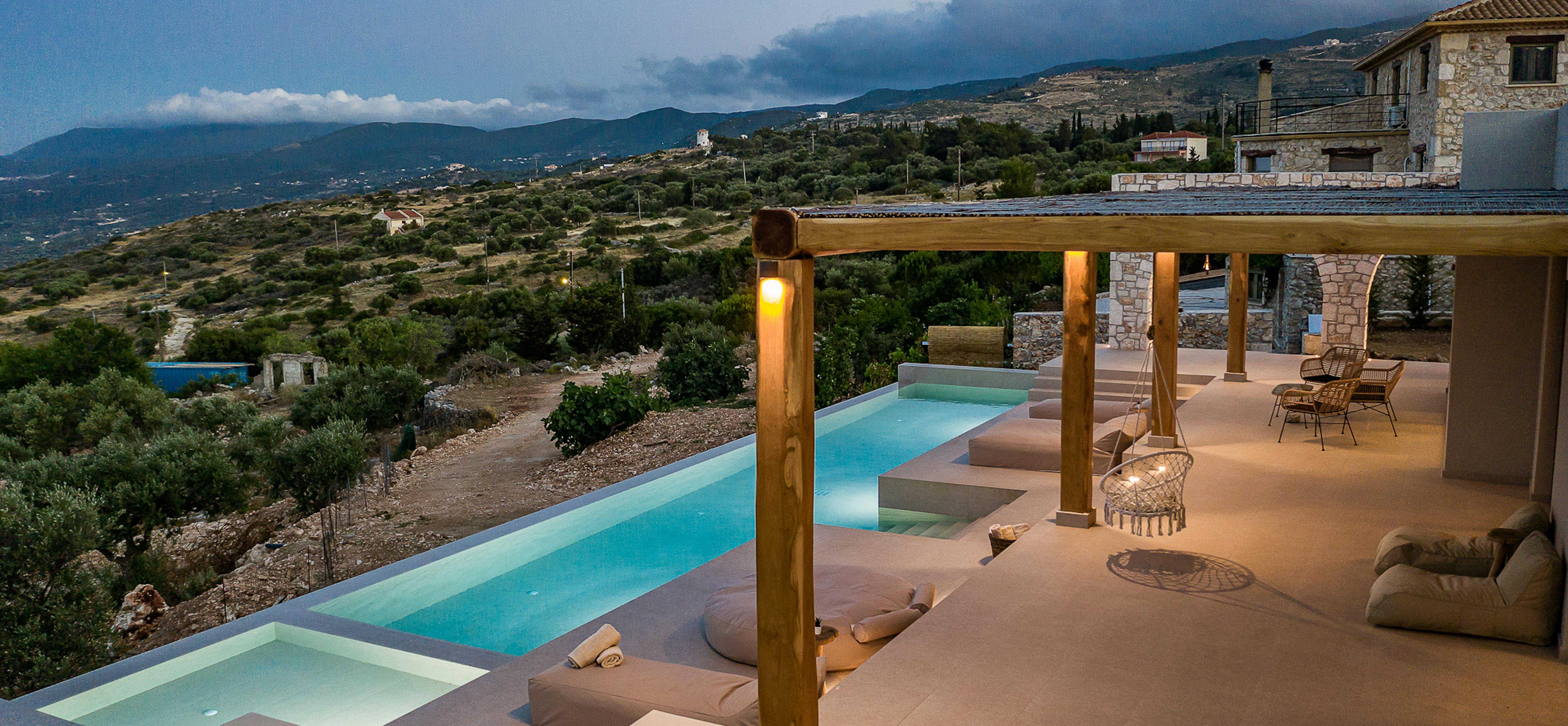 Illuminated infinity pool terrace at dusk with hot tub, wooden pergola, and mountain valley views.