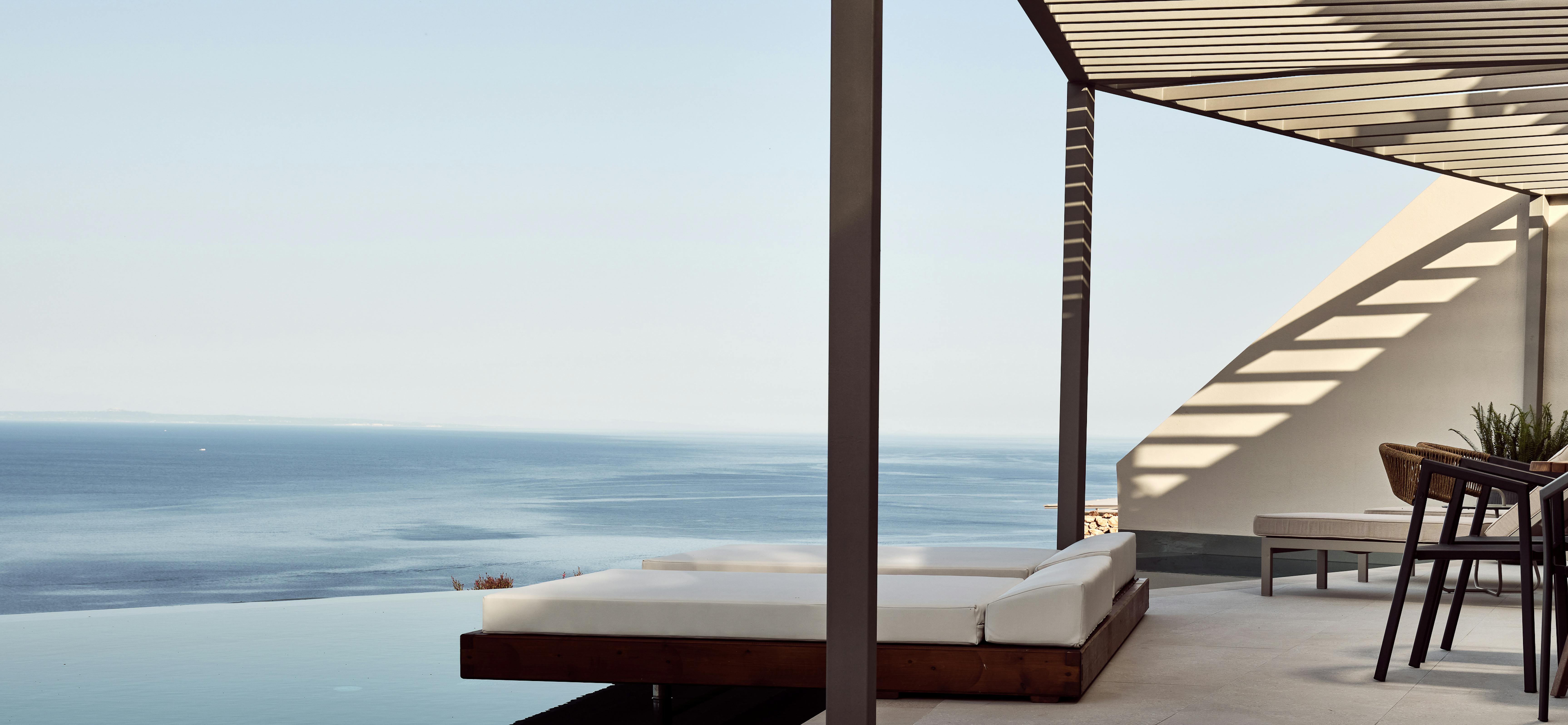 Pool terrace with white cushioned loungers under pergola overlooking coastal horizon.