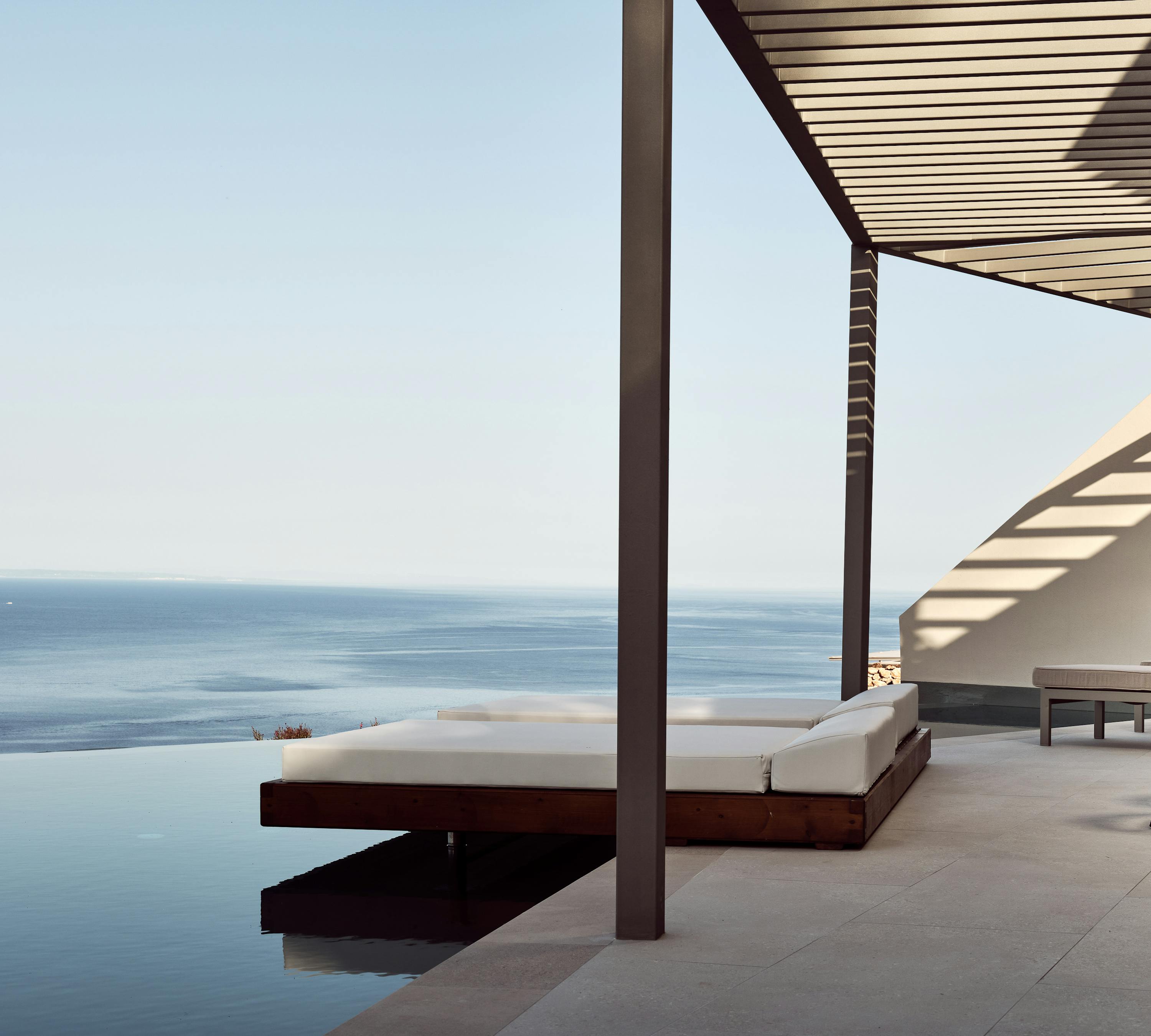 Pool terrace with white cushioned loungers under pergola overlooking coastal horizon.