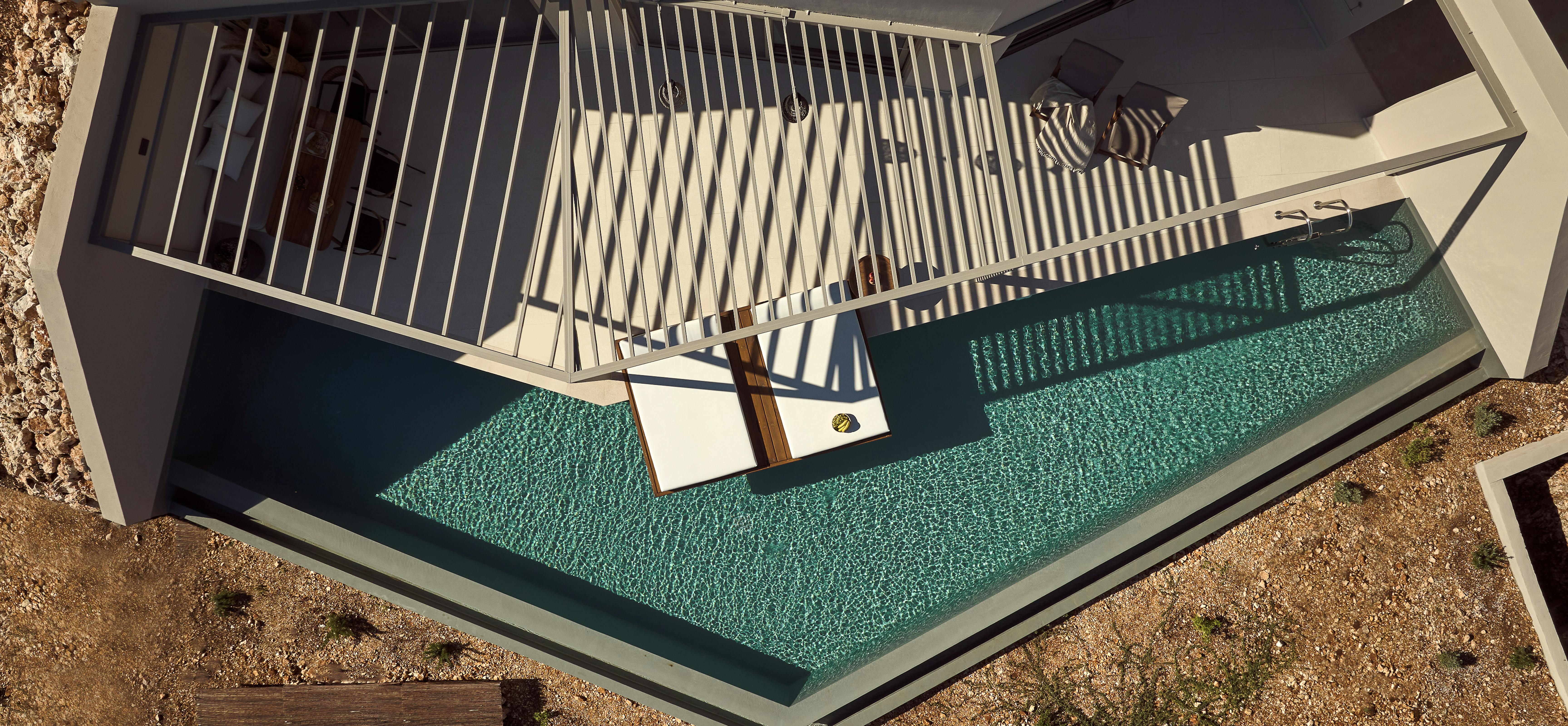 Aerial view of angular infinity pool with floating daybed and geometric white pergola.