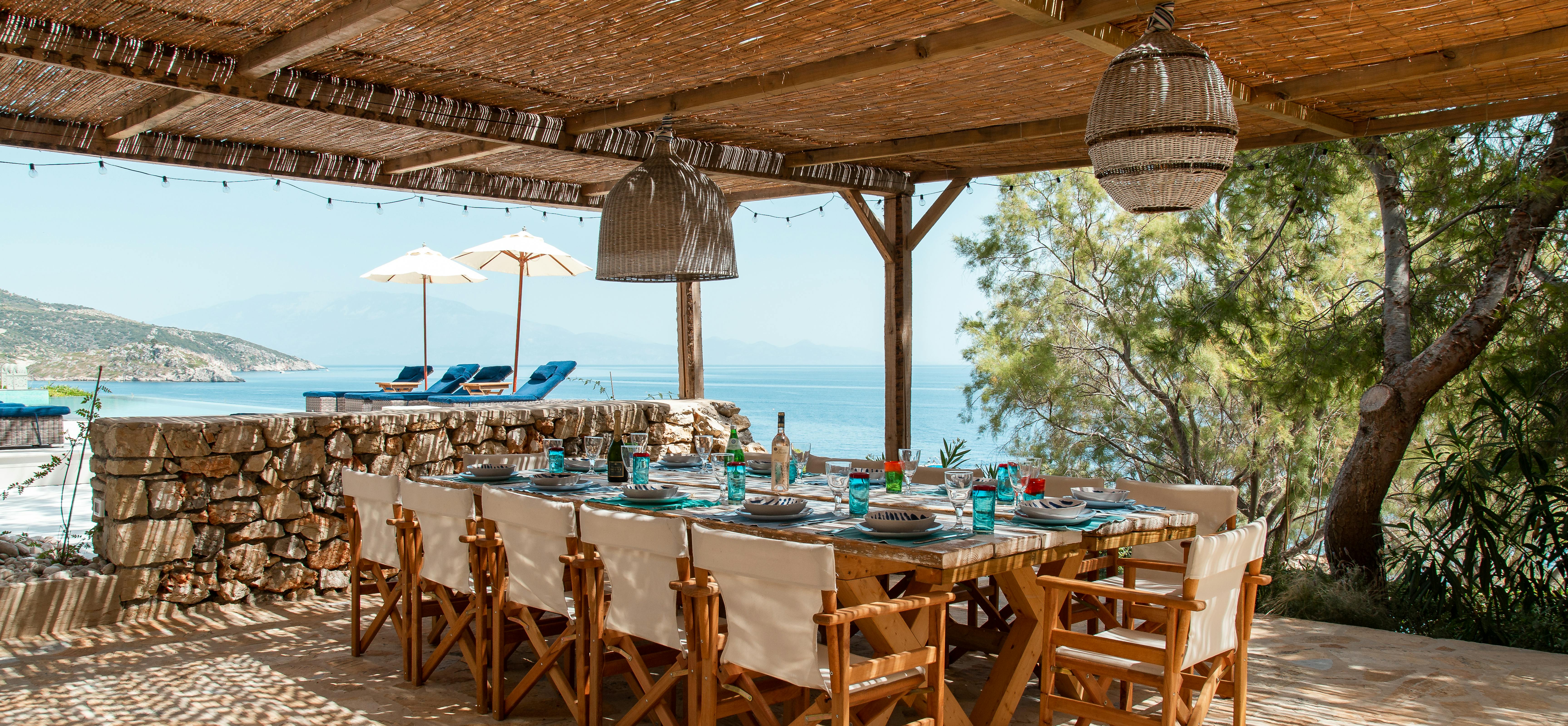 Covered outdoor dining terrace with bamboo ceiling and wooden director's chairs around long table. Stone wall and blue water views create Mediterranean coastal dining atmosphere.