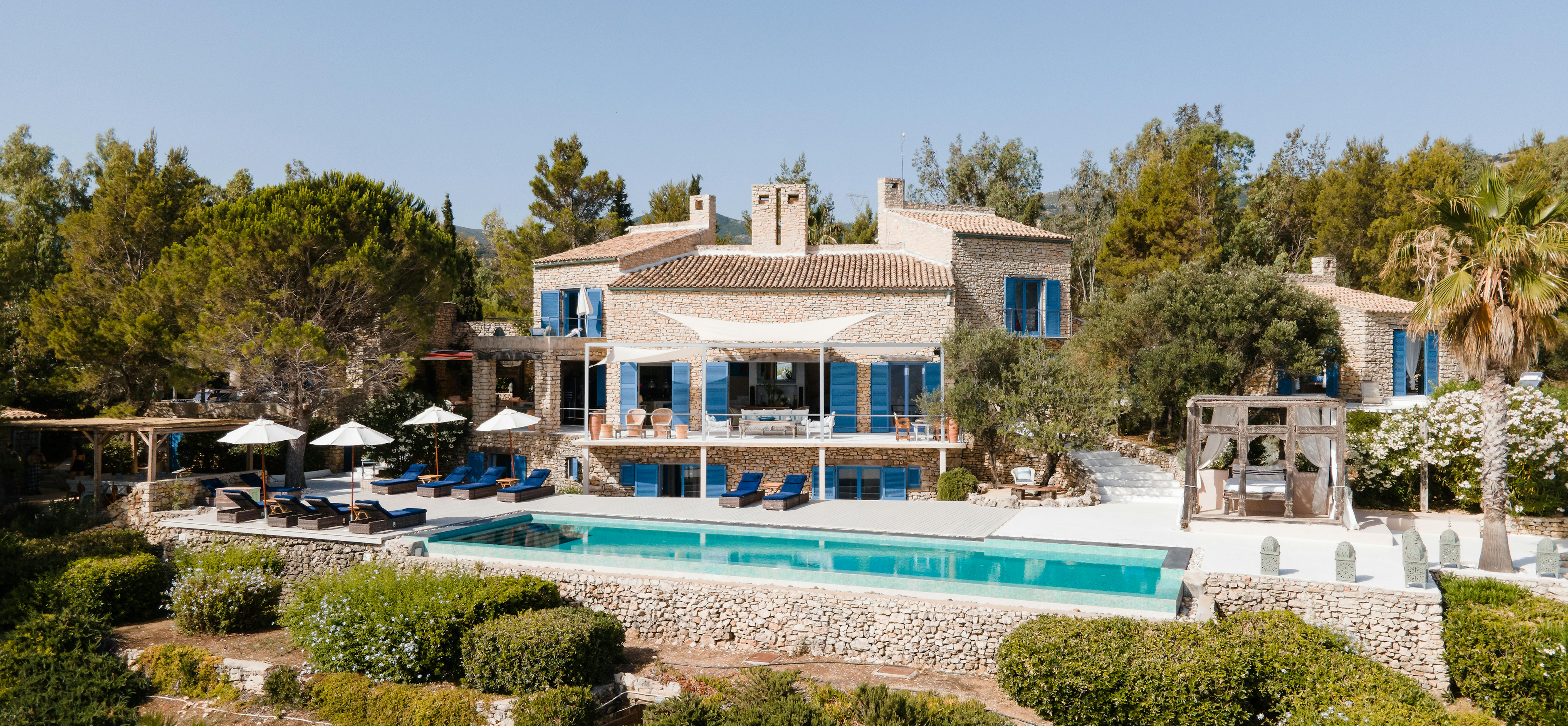 Aerial view of stone Mediterranean villa with blue shutters and large swimming pool. Multi-level terraced property surrounded by lush gardens and natural vegetation.