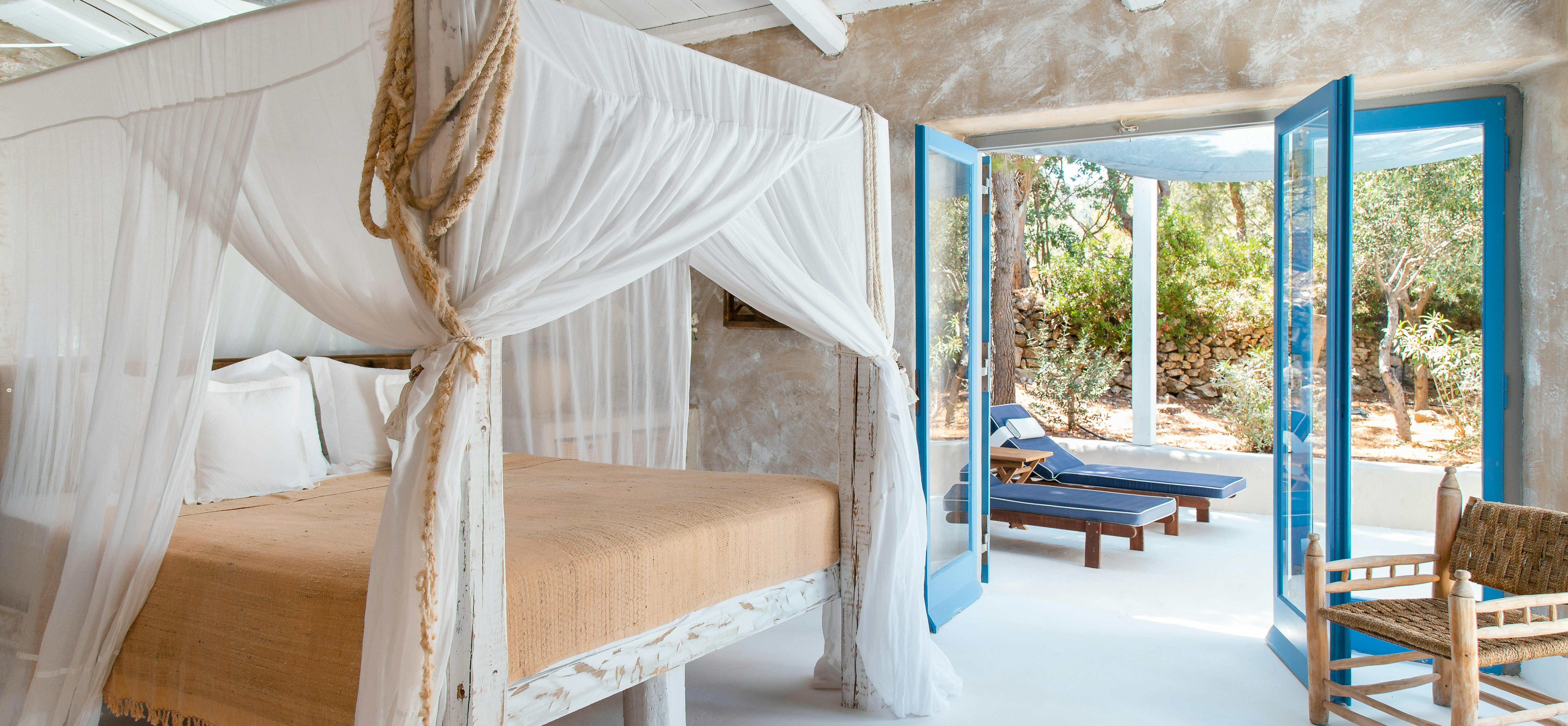 Rustic bedroom with white four-poster bed and mosquito netting under exposed beam ceiling. Blue French doors open to terrace with outdoor loungers and natural landscape views.