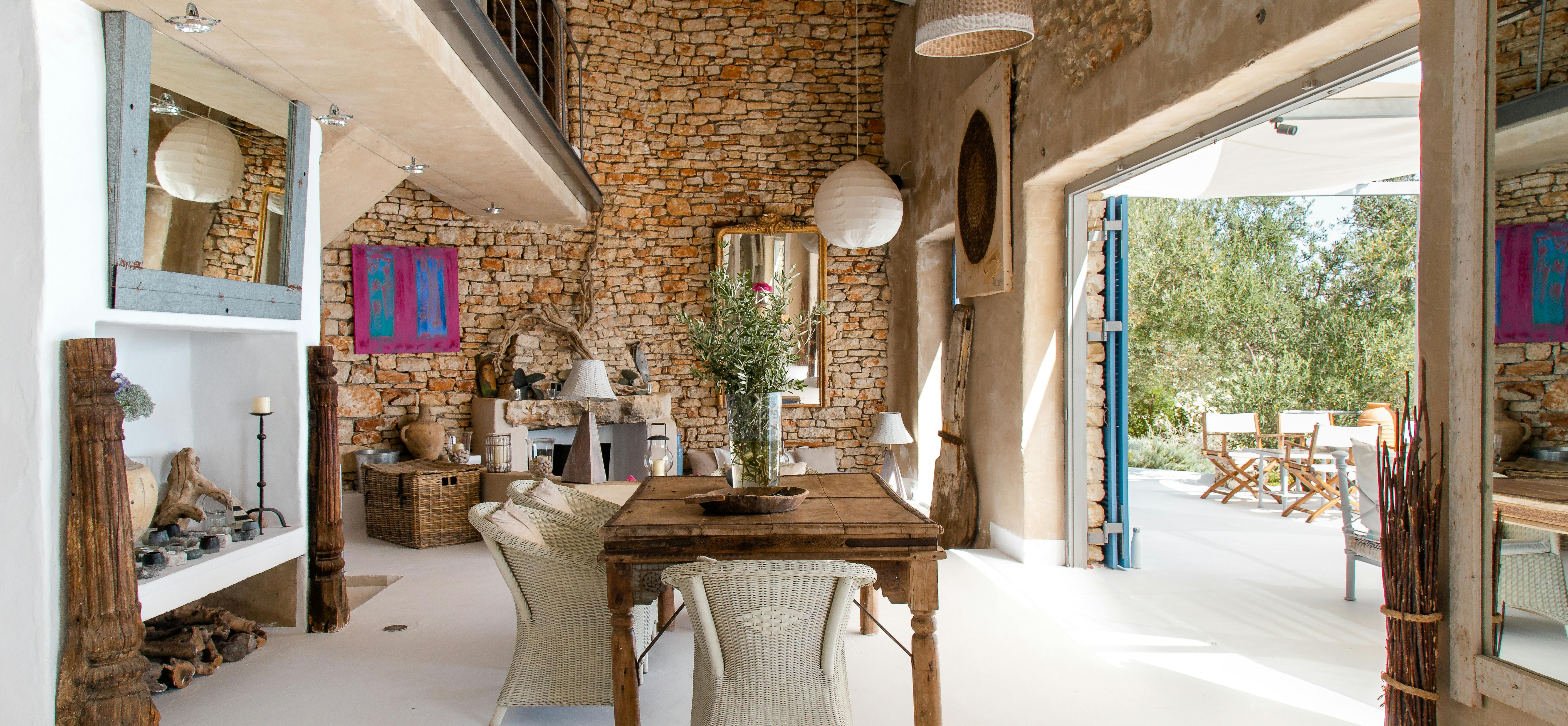 Open-plan living space with exposed stone walls and rustic wooden dining table. White built-in shelving and wicker furniture complement traditional Mediterranean architecture.