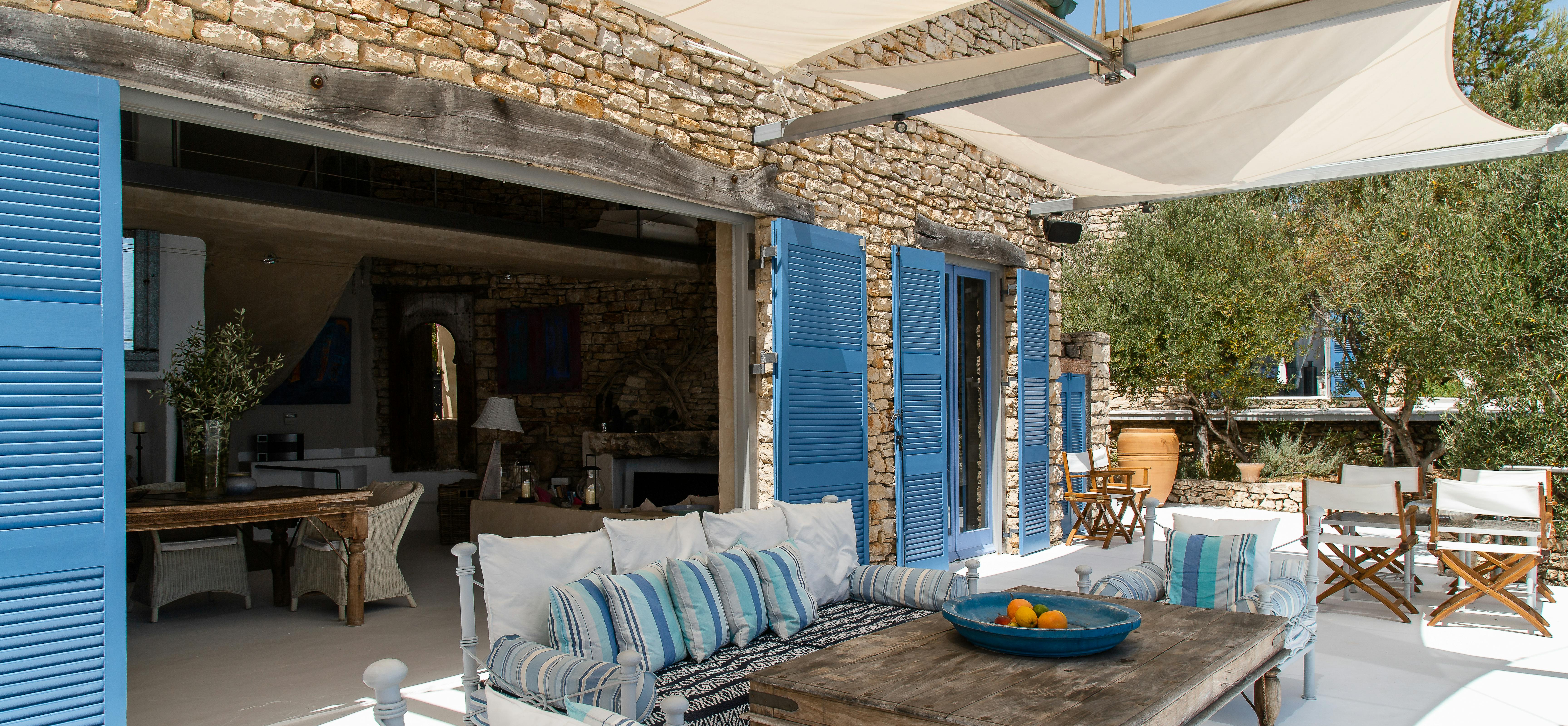 Covered outdoor living area with white furniture and blue striped cushions. Stone building with blue shutters forms backdrop to shaded terrace.