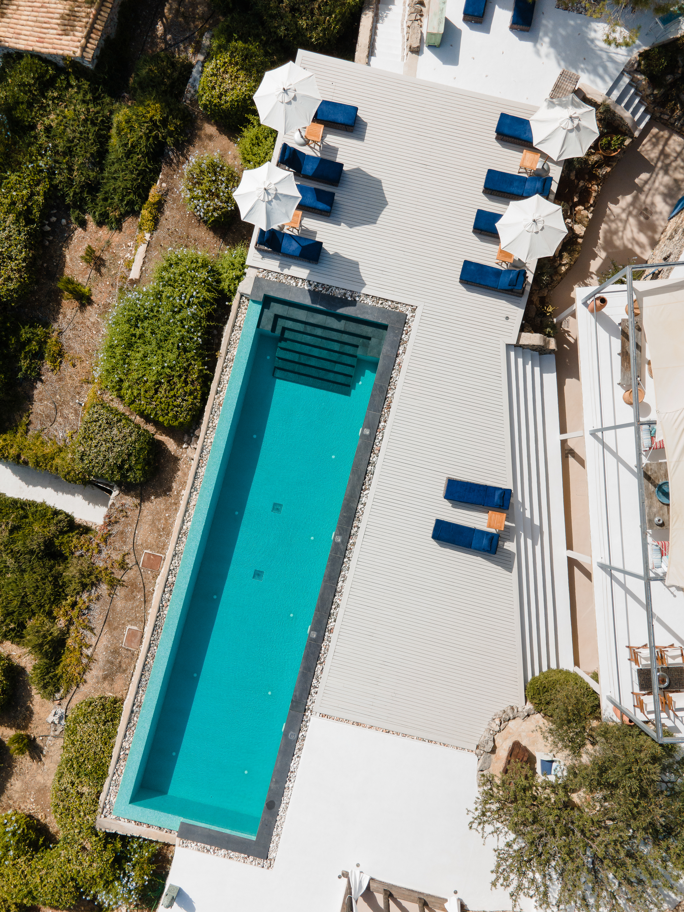 Aerial view of rectangular swimming pool with blue loungers arranged on white decking. Property surrounded by natural Mediterranean landscape and olive trees.