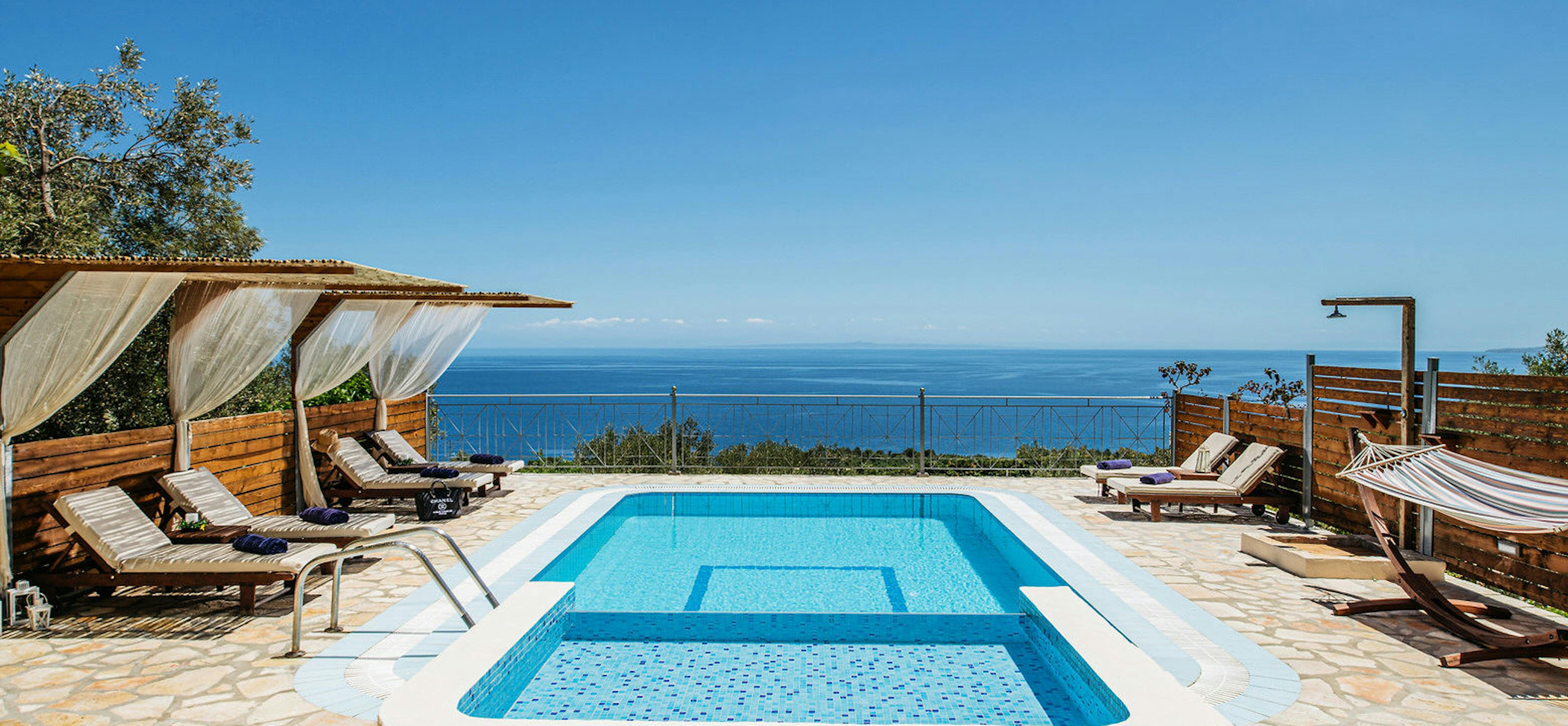Luxury Mediterranean pool terrace with elevated spa section, wooden loungers, and panoramic ocean views under clear blue skies.