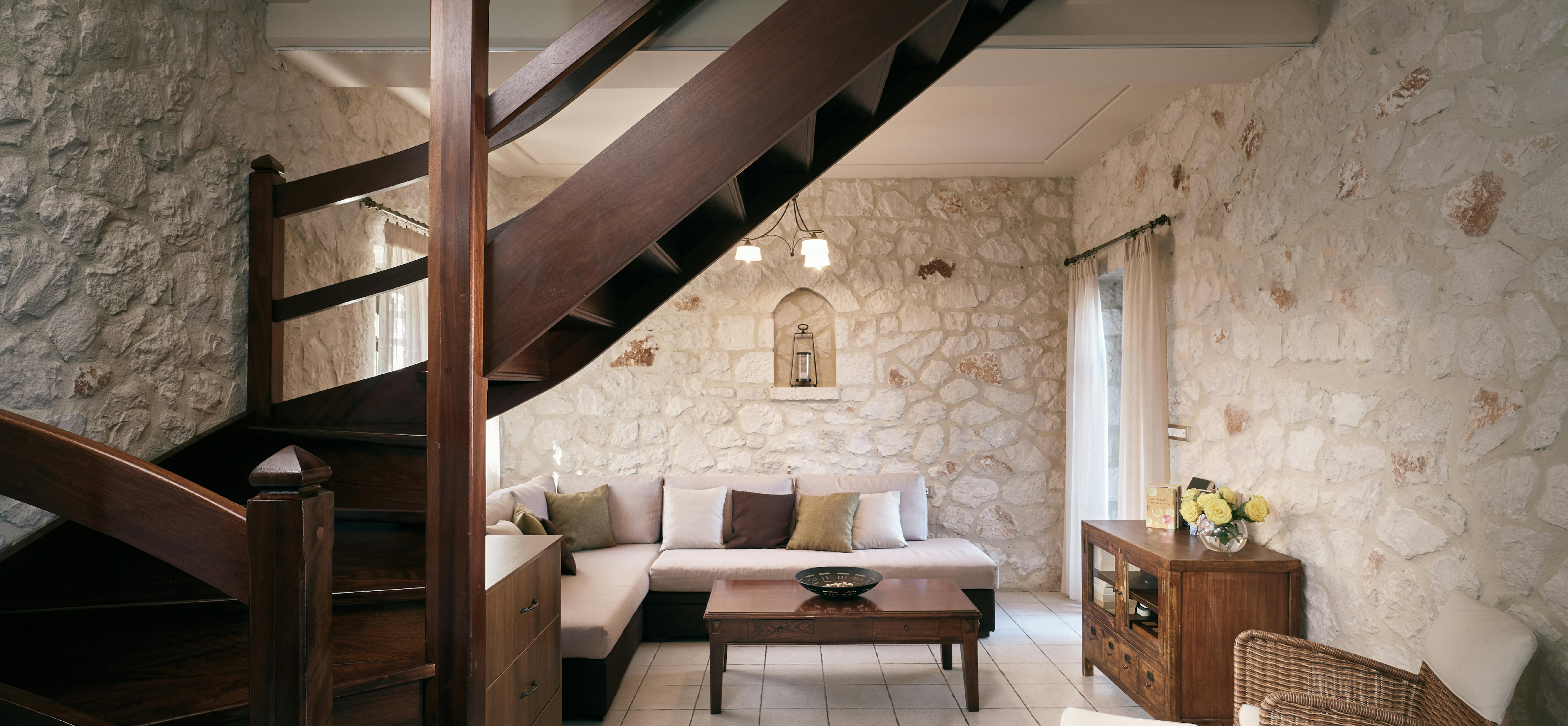 Rustic living room with wooden staircase, stone walls, and neutral-toned sectional sofa under exposed beams.