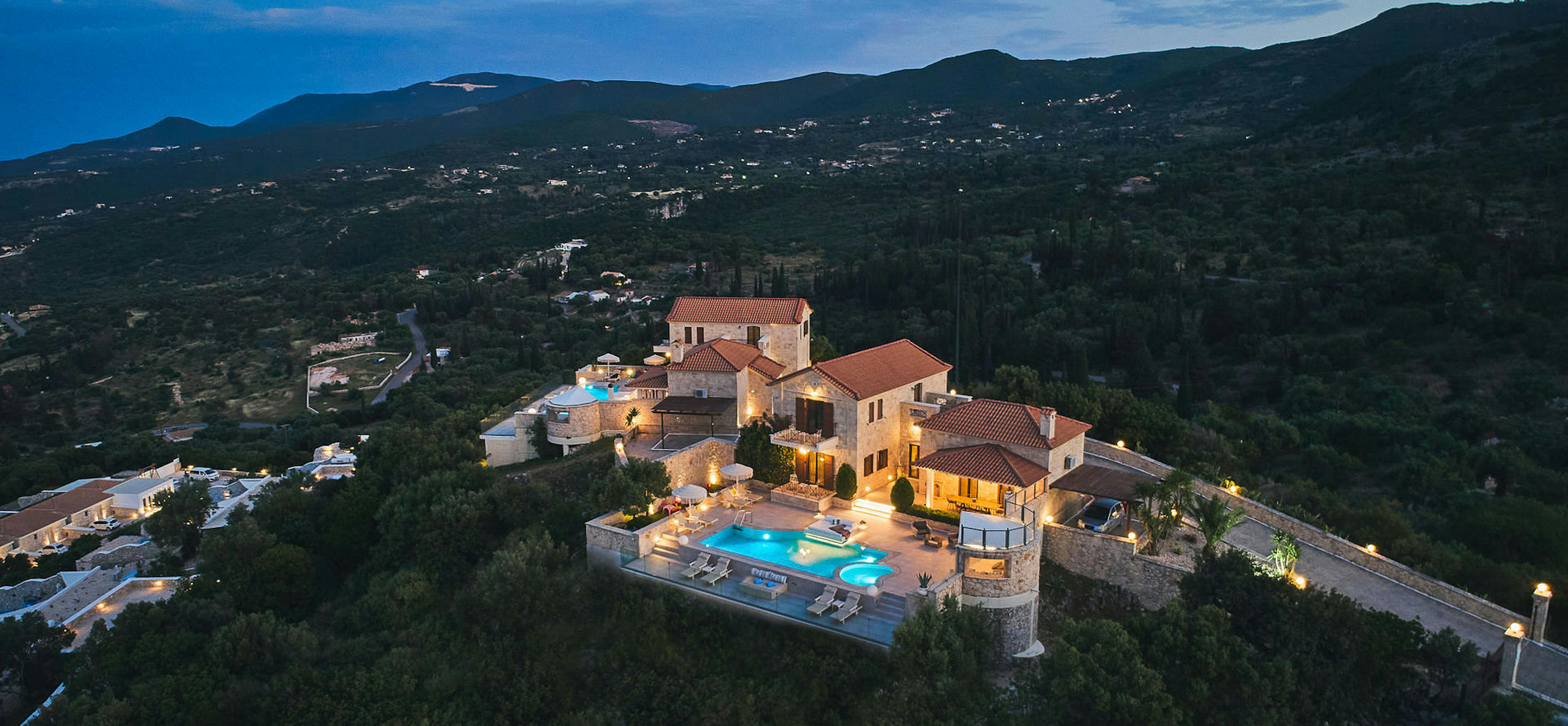 Aerial view of Mediterranean villa complex with terracotta roofs, pools, and mountainous landscape backdrop.