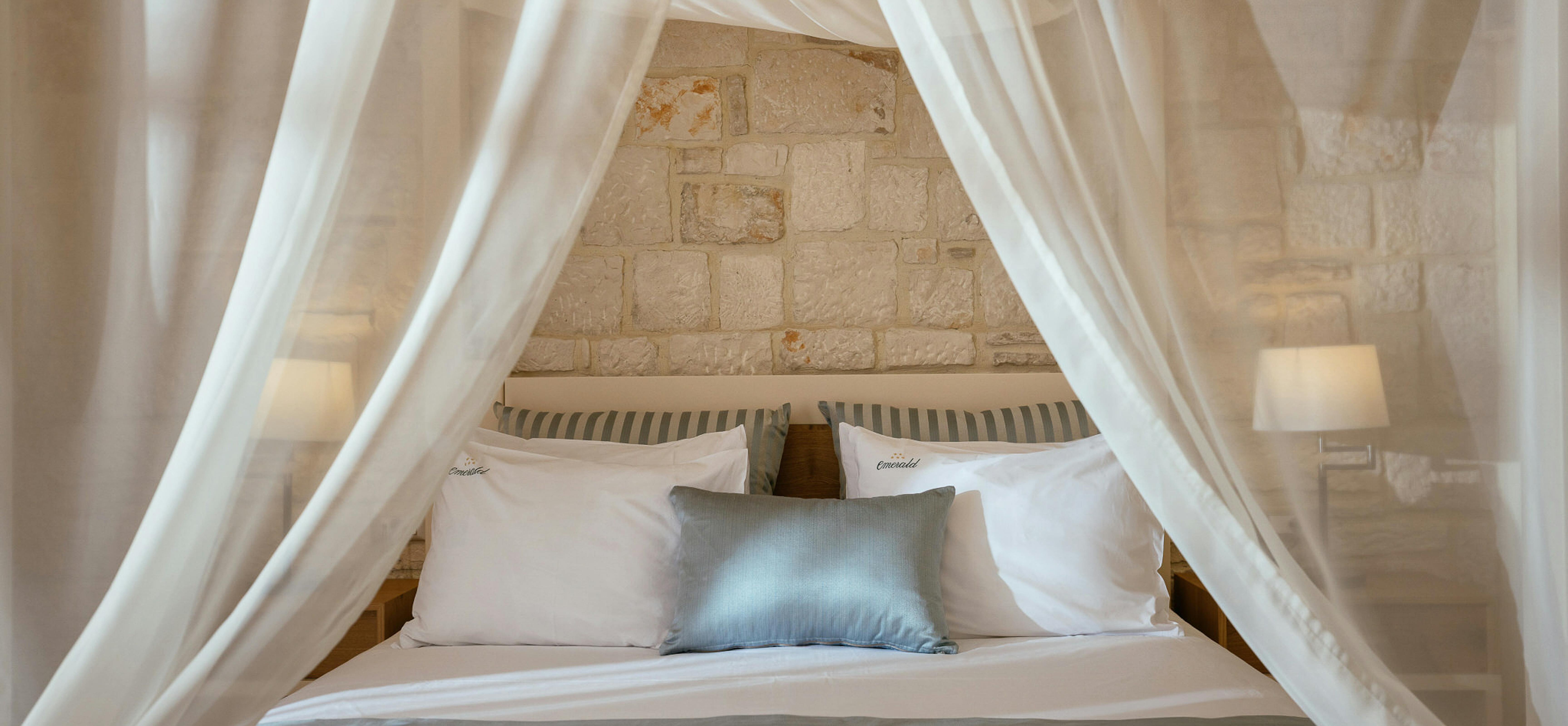 Romantic canopy bed with flowing white curtains, stone walls, and soft blue accent pillows.