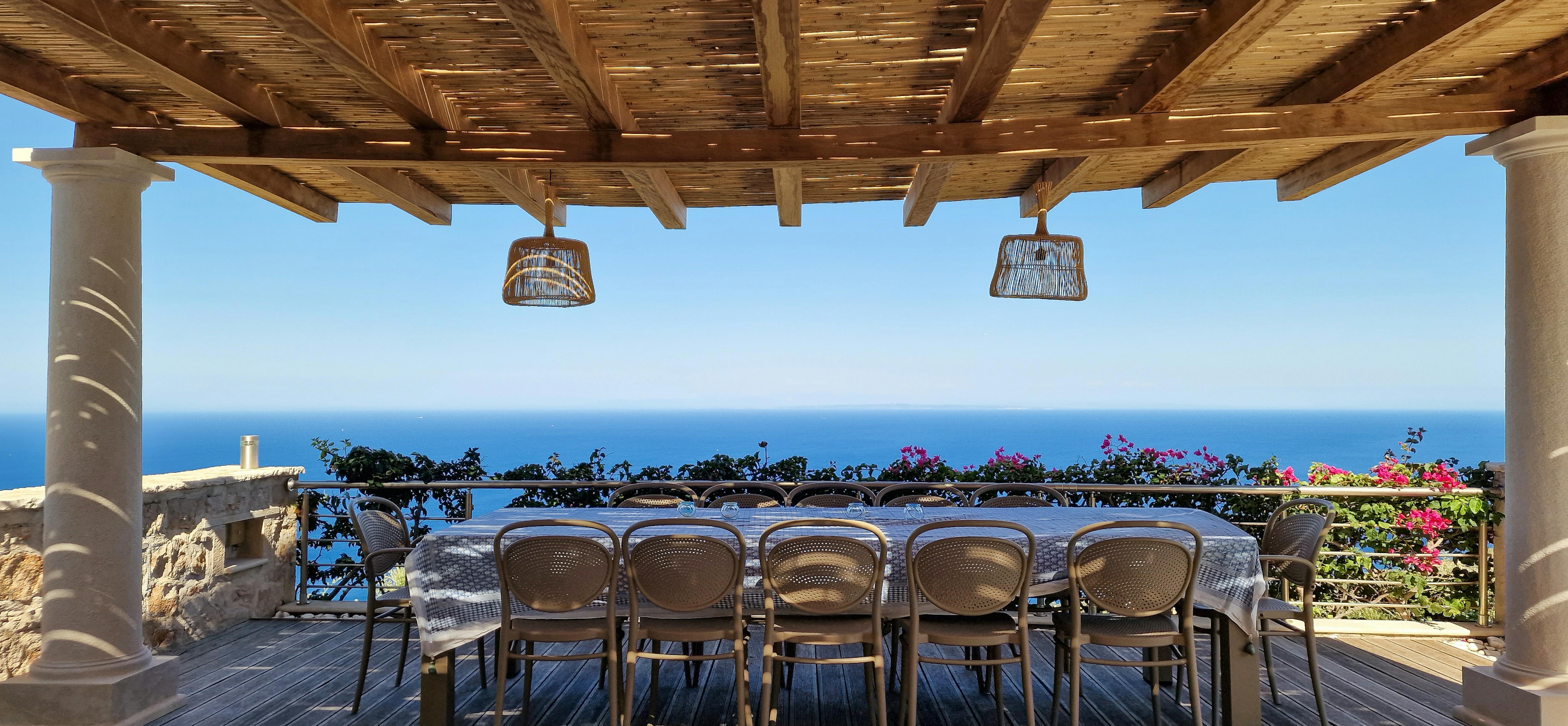 Covered dining terrace with long table, wicker pendant lights, and panoramic ocean backdrop.
